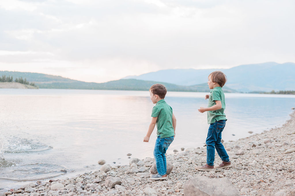 Two toddler brothers in matching jeans and green polos throw rocks in an alpine lake at sunset after finding Best Places to Eat with Kids in Breckenridge