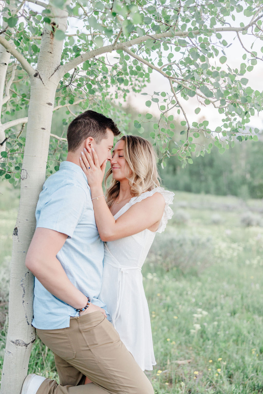 A woman in a white dress holds the cheeks of her partner leaning on a tree in a mountain meadow during a romantic getaway breckenridge