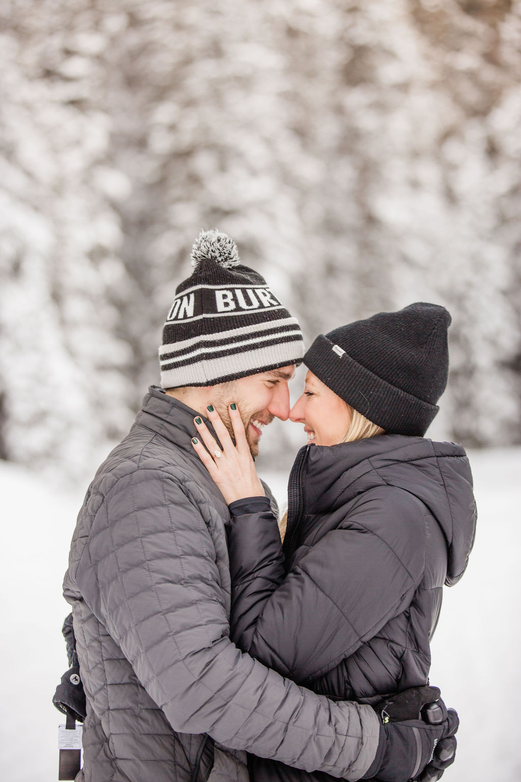 A happy couple touches noses in the snow in black jackets and beanies