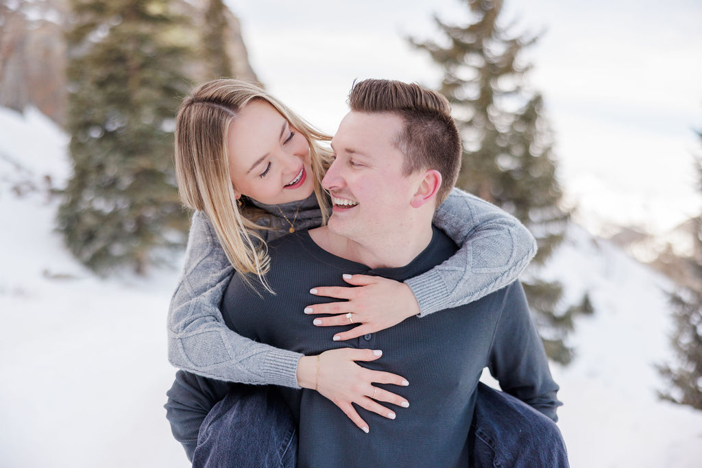 A woman climbs on the back of her fiancee as they play on a snowy hill at sunset in the mountains