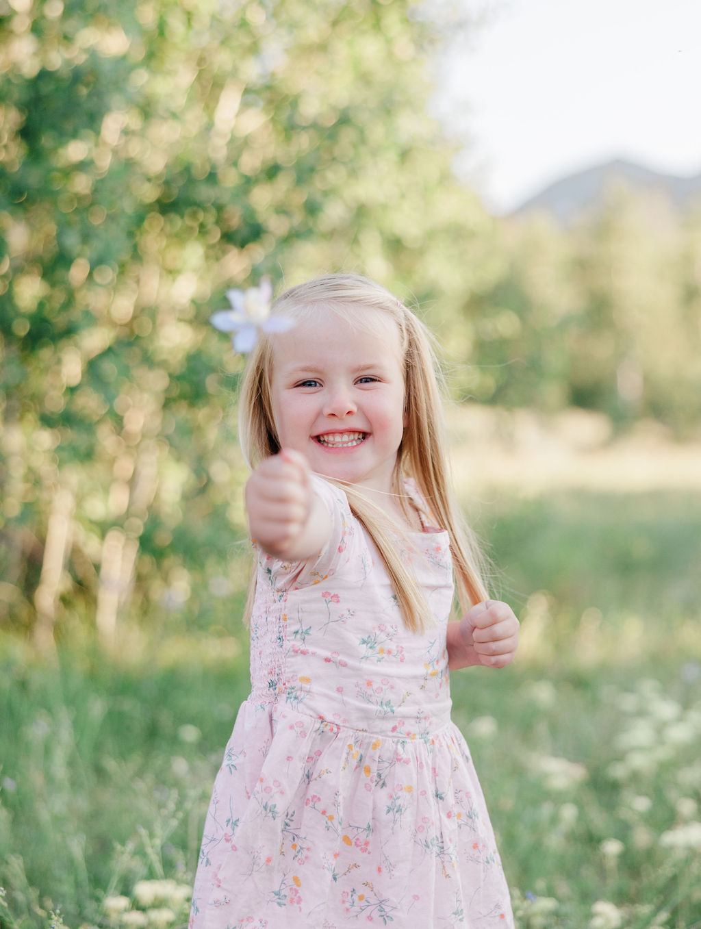 A happy toddler girl in a pink dress shows off a flower in a meadow at sunset