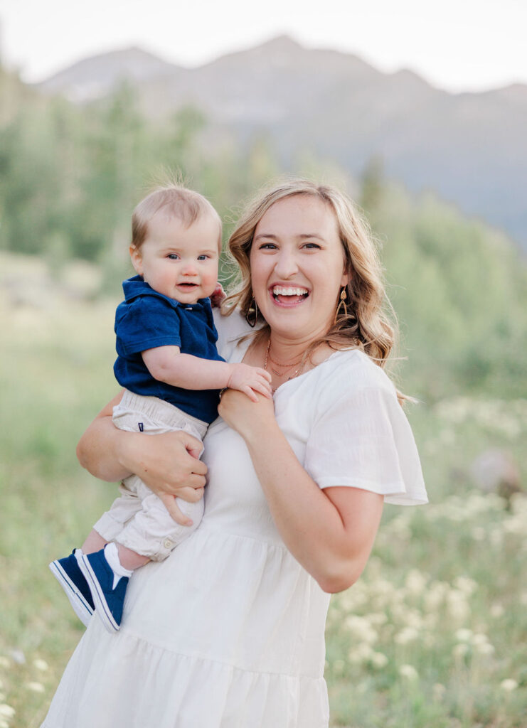 A happy mom laughs in a white dress while standing in a meadow playing with her toddler son on her hip during summer family activities breckenridge