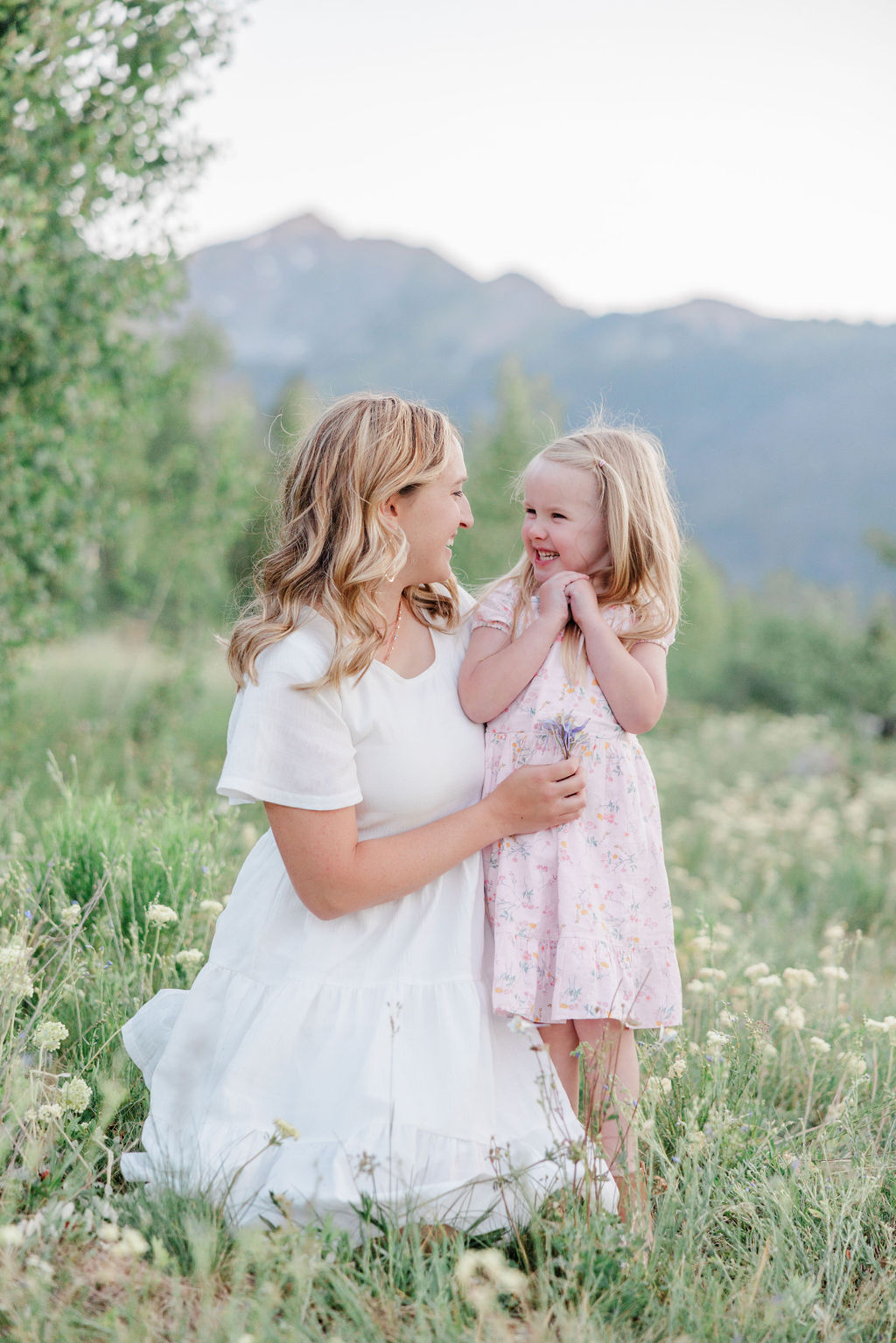 A happy toddler giggles while playing with mom in a pink and white dress in a meadow during summer family activities breckenridge