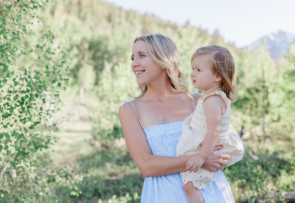 A mom and her toddler daughter on her hip explore a mountain trail with smiles