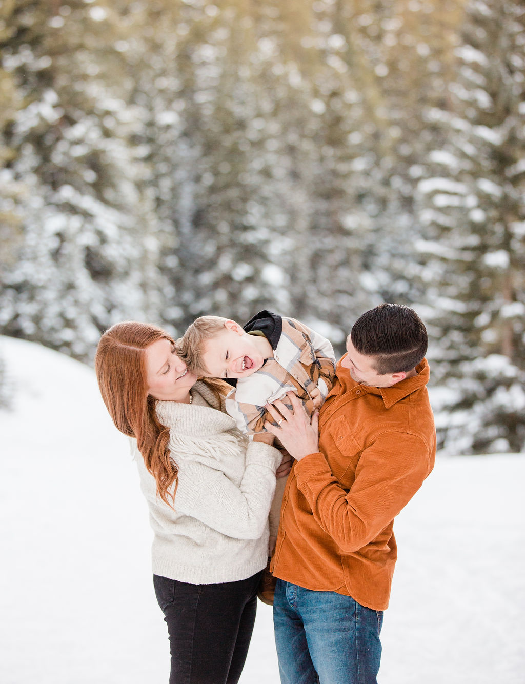 A happy toddler in plaid is tickled while being held by mom and dad in a mountain trail in snow after meeting breckenridge nannies