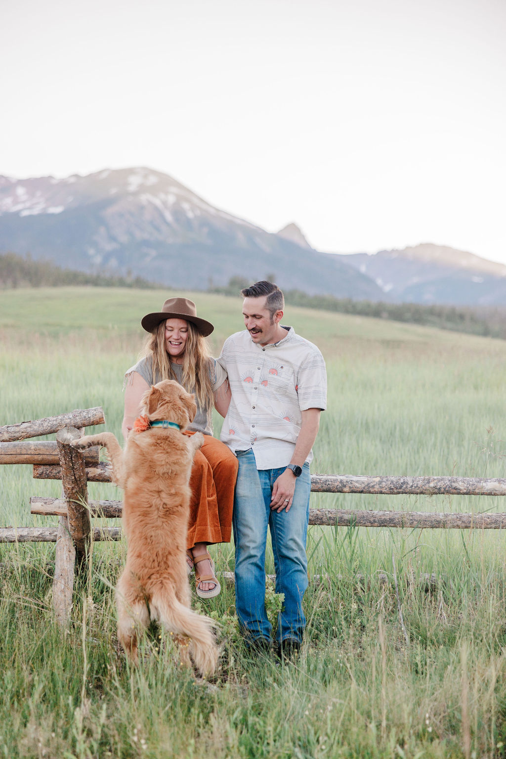 A couple sits on a wooden fence in a large pasture playing with their golden retriever