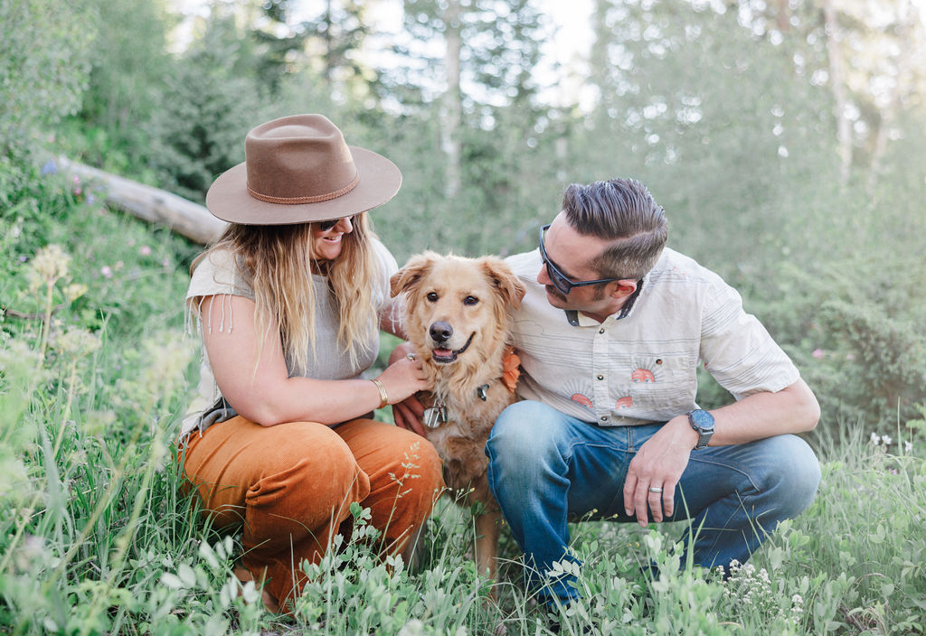 A smiling couple in white shirts squat with their happy dog in a mountain meadow during one of the date ideas in breckenridge