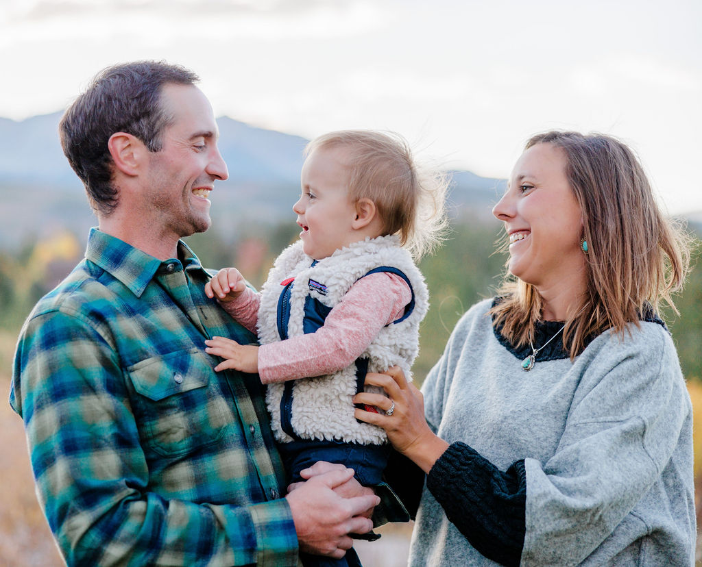 A happy baby girl sits in dad's arms while playing in a park with mom and dad after visiting a daycare in breckenridge