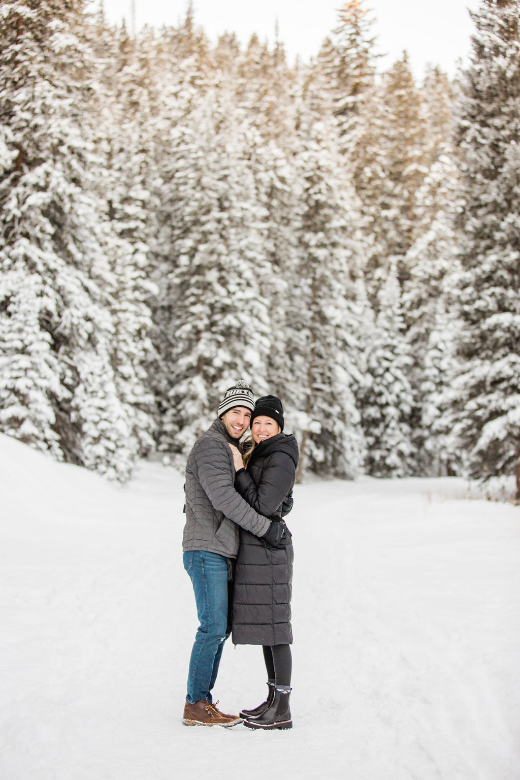 A happy engaged couple hugs and smiles while snuggling on a snow covered mountain trail