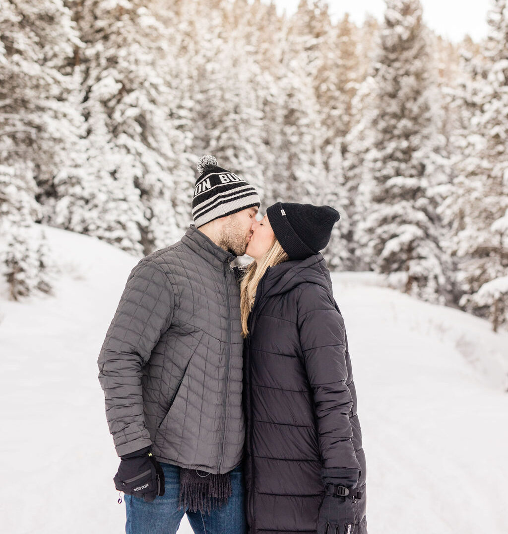 A newly engaged couple kisses in snow covered mountains in thick black coats