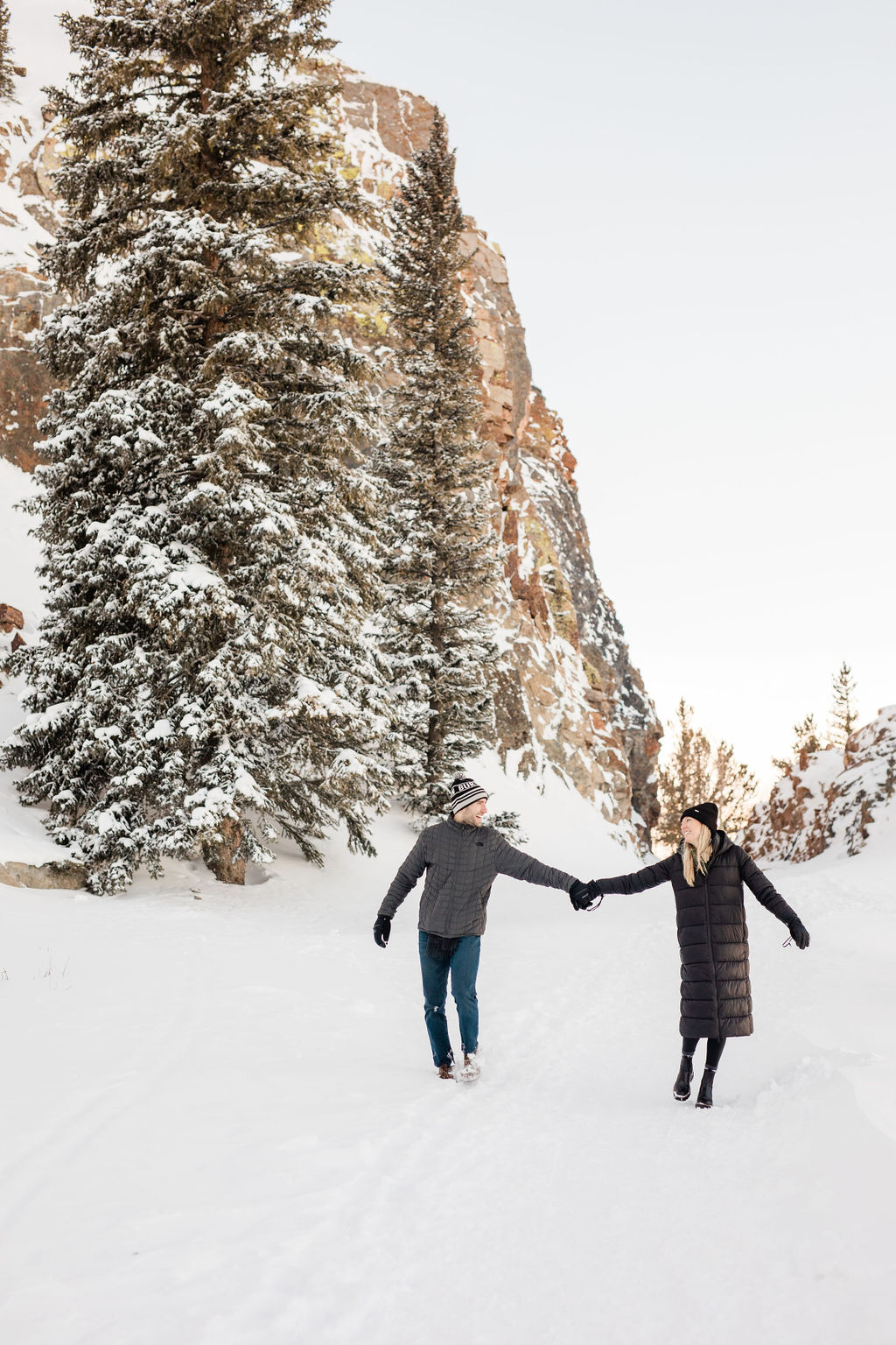 A laughing engaged couple holds hands and plays in a mountain trail covered in snow in large coats after using jewelry stores in breckenridge colorado