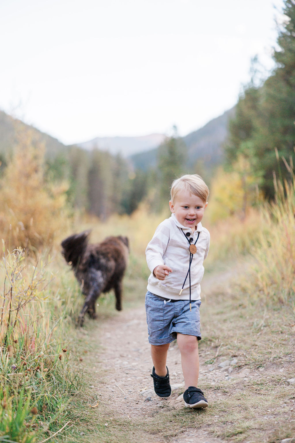 A toddler boy in a bolo tie and white shirt runs in a trail with a dog before visiting playgrounds in breckenridge