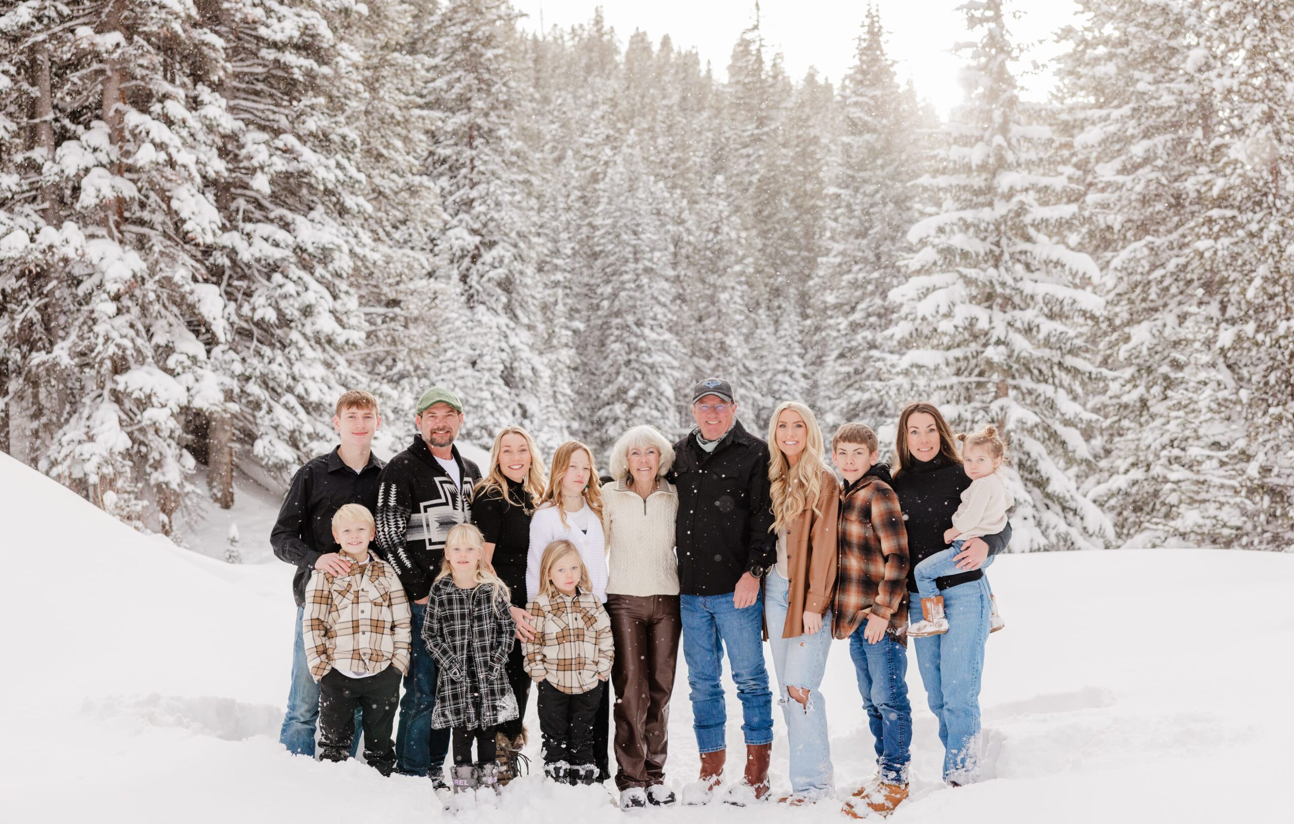 An extended family of 14 people are standing close together and smiling in a wintery scene of tall pine trees covered in snow.