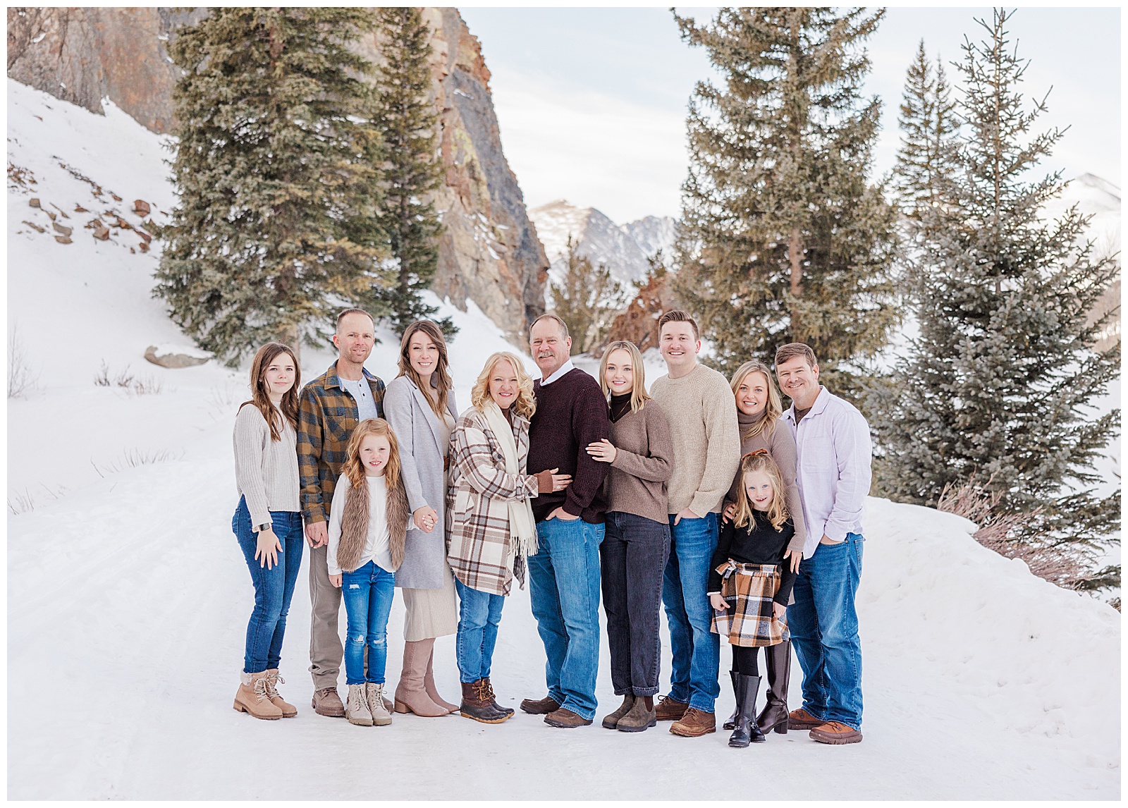 An extended family of 12 people are standing close together, smiling at the camera. A winter scene of pine trees, tall cliffs, and snow are behind them.