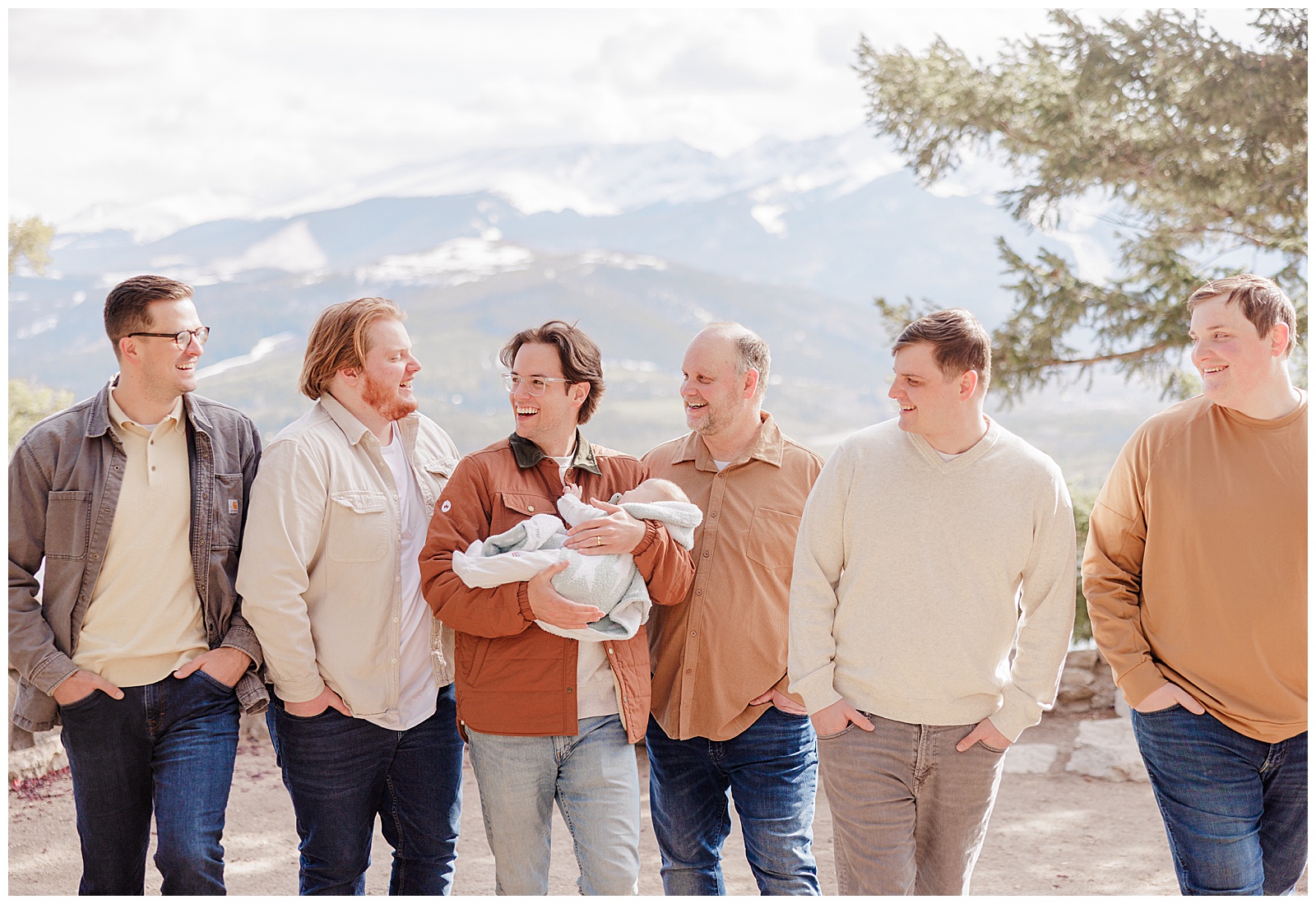 This is photo of 6 adult men, one with their newborn child. They are all casually walking and smiling with each other. There is a mountain scene behind them. 