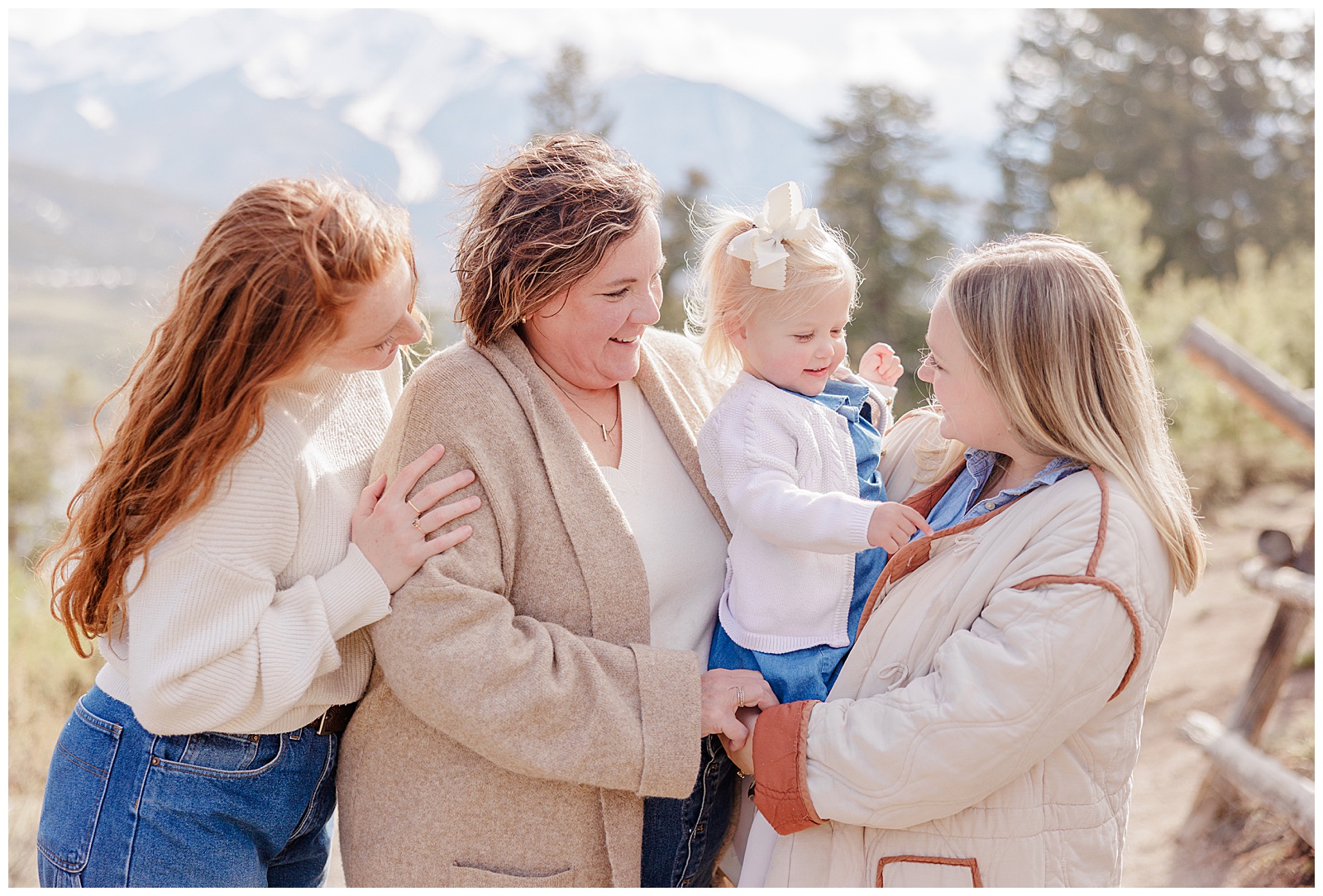 In this photo there are 3 adult women with one small child. They are holding her and all smiling with one another. There is a mountain with pine trees behind them. 