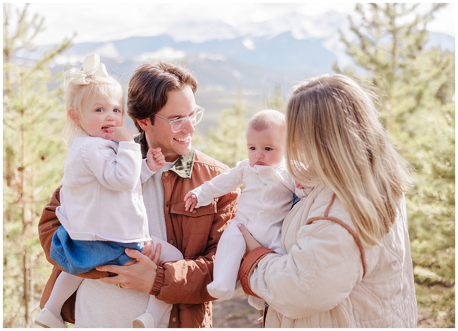 This is a family of four with two adults and two young children. The parents are holding their children in their arms. They are all smiling at one another, with gorgeous mountains and trees behind them. 