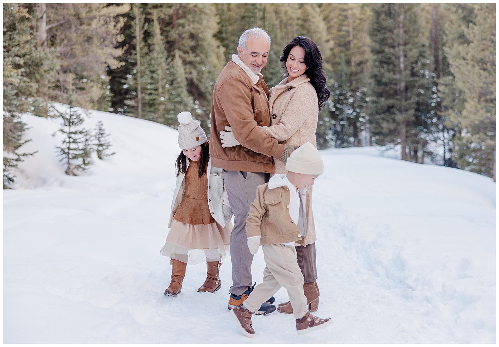 This is a photo of a family of four. The mom and dad are huddled close together. Their two young children are running playfully around them. They are surrounded by gorgeous pine trees covered in snow. Shot by Breckenridge Photographer, Rachel. 