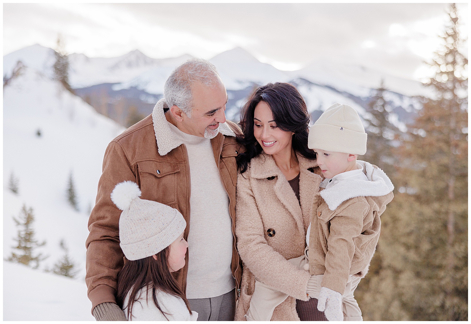 This is a family of 4. They are standing closely together, smiling at one another. There are gorgeous snow covered peaks behind them. Shot by a Breckenridge family photographer.