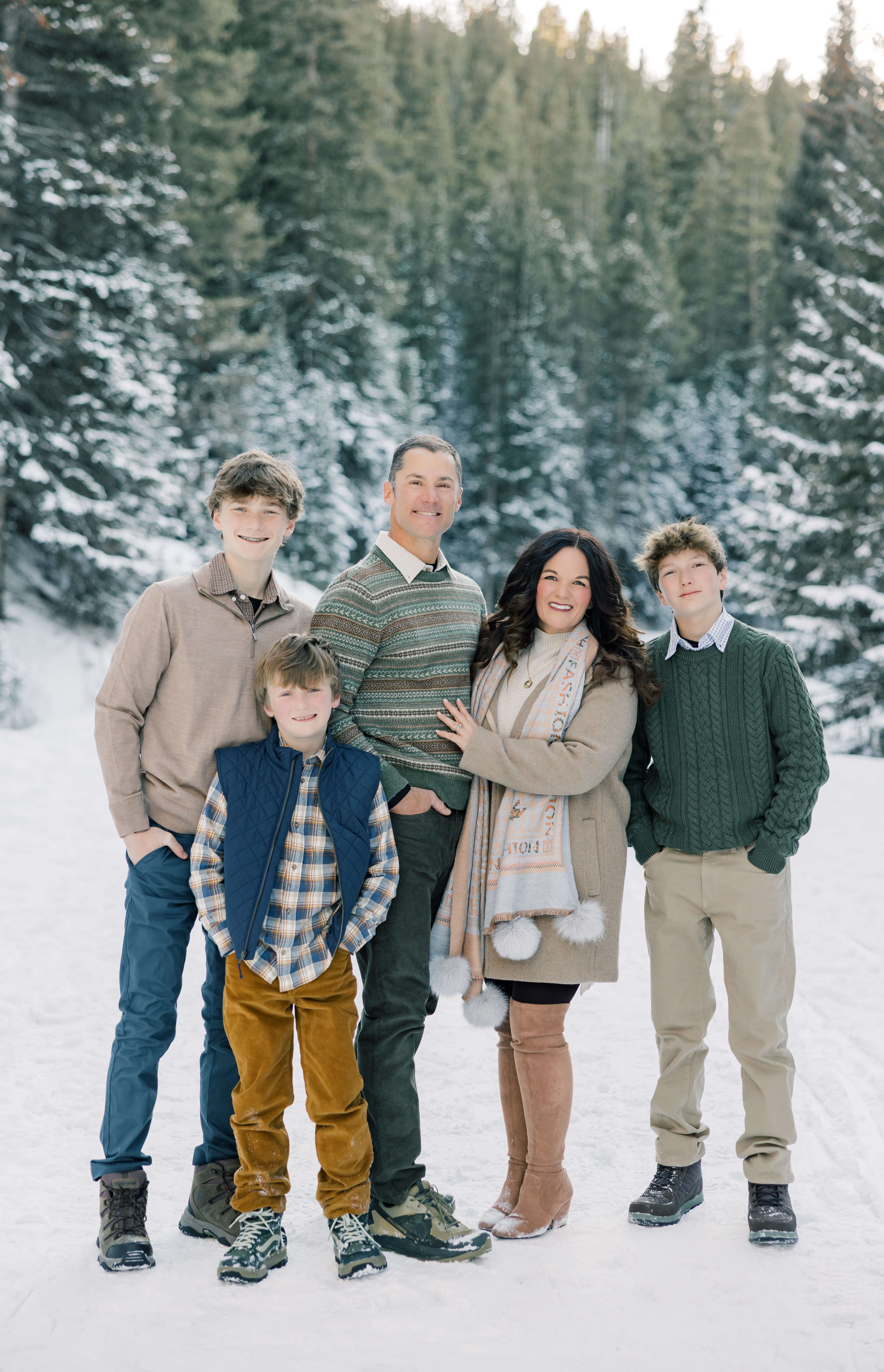 A mom and dad stand with their three teen sons in a snow covered trail in a forest at sunset