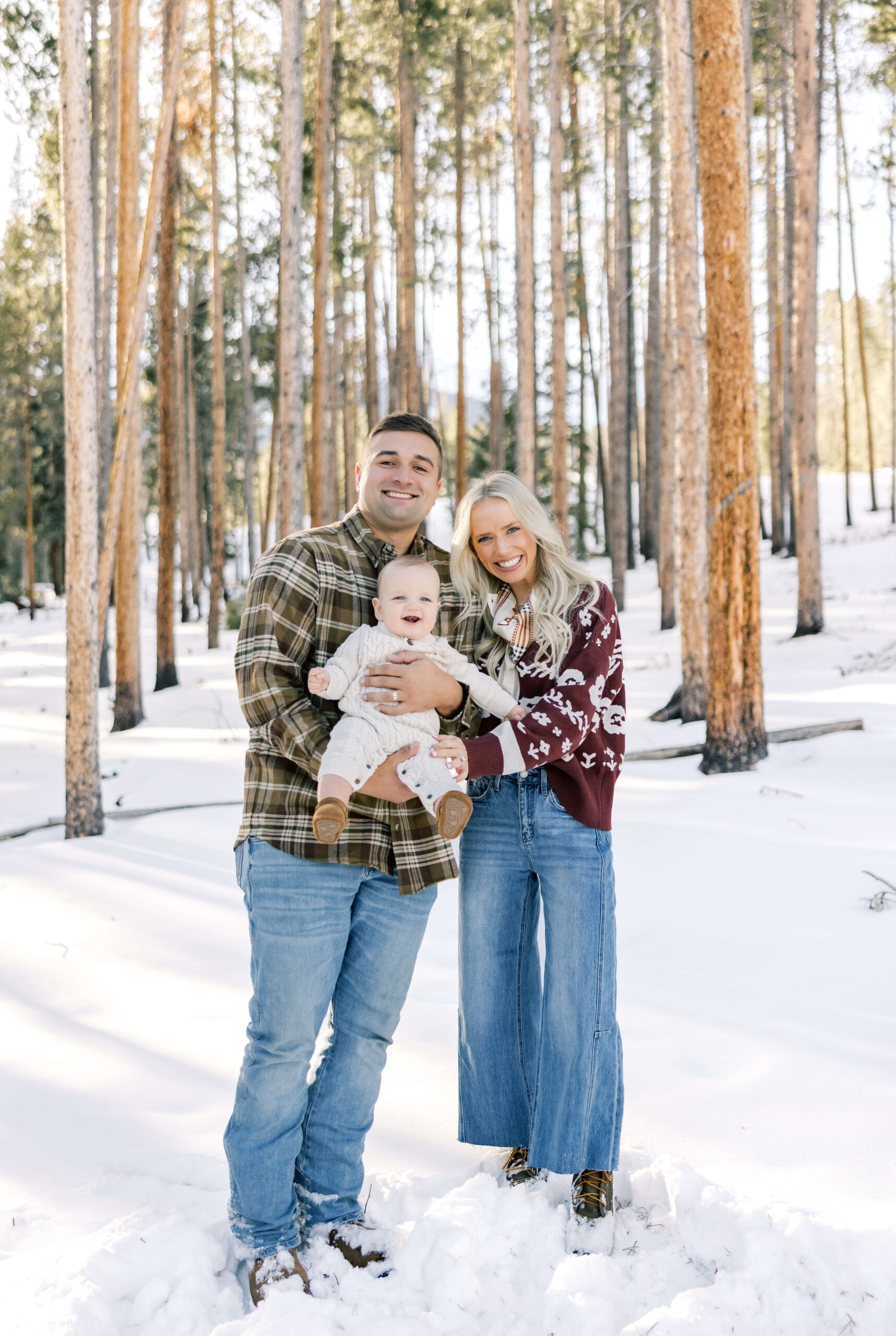 Happy mom and dad stand in a snow covered forest in jeans and sweaters holding their happy baby