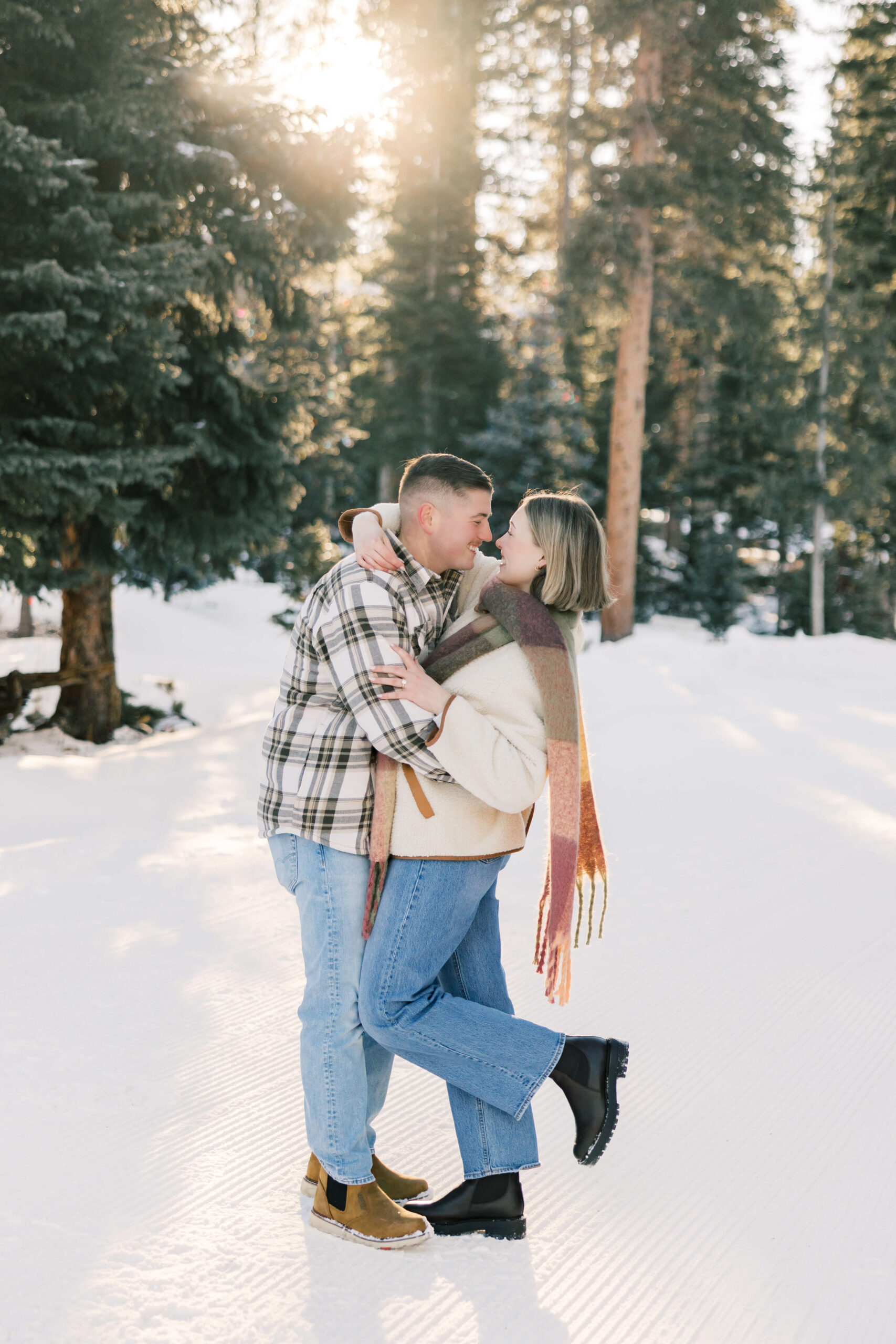 A happy couple embrace and lean in for a kiss in a snow covered forest at sunset in plaid before some Fine Dining in Breckenridge CO