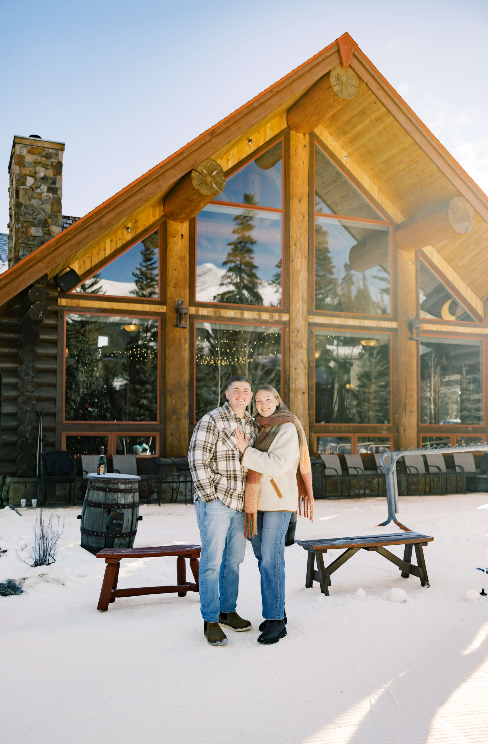 A happy newly engaged couple smile while standing and snuggling behind a snow covered cabin before enjoying some Fine Dining in Breckenridge CO