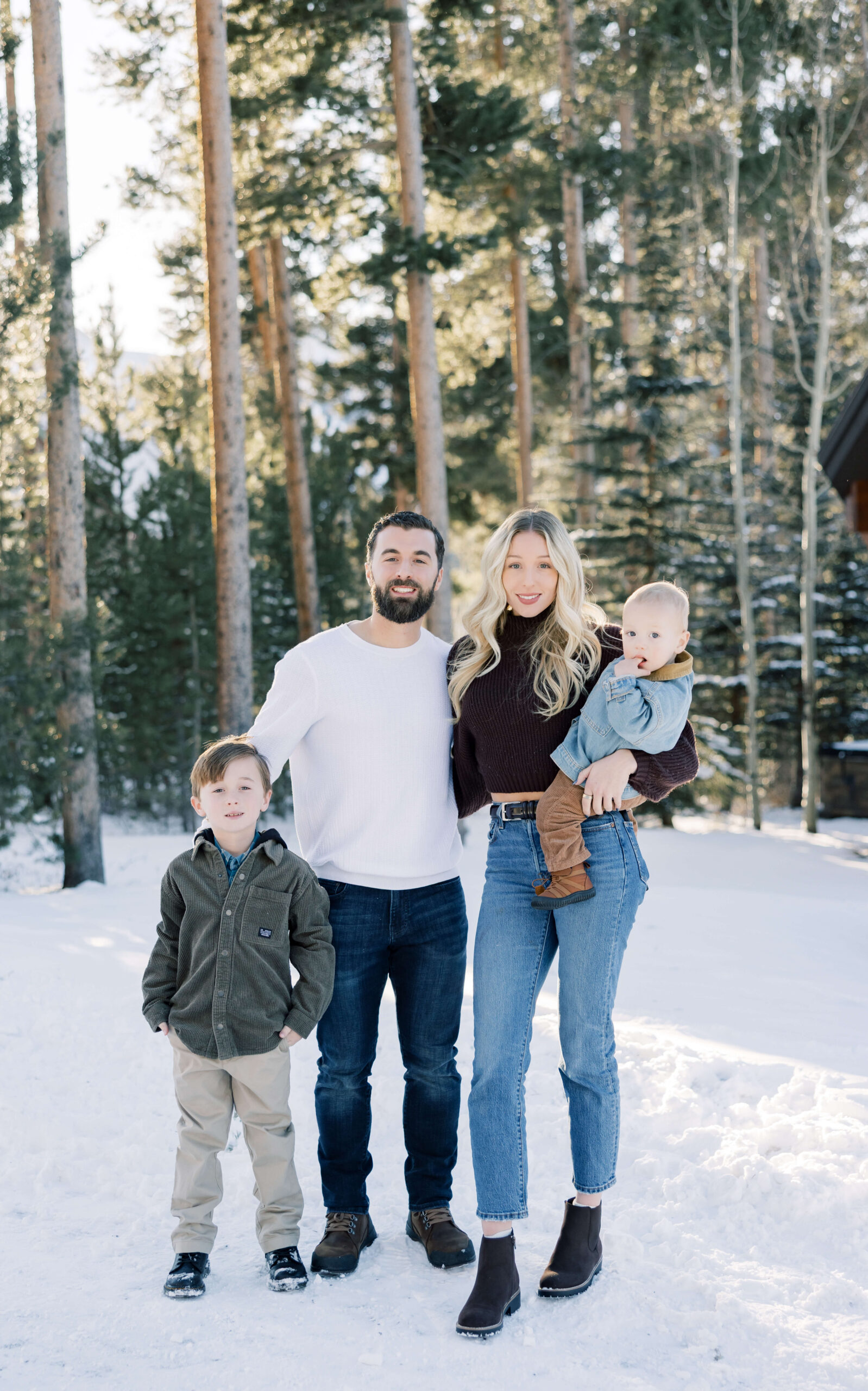 A mom and dad stand in a snow covered forest holding a toddler and an arm around their young son before some Snow Tubing in Breckenridge