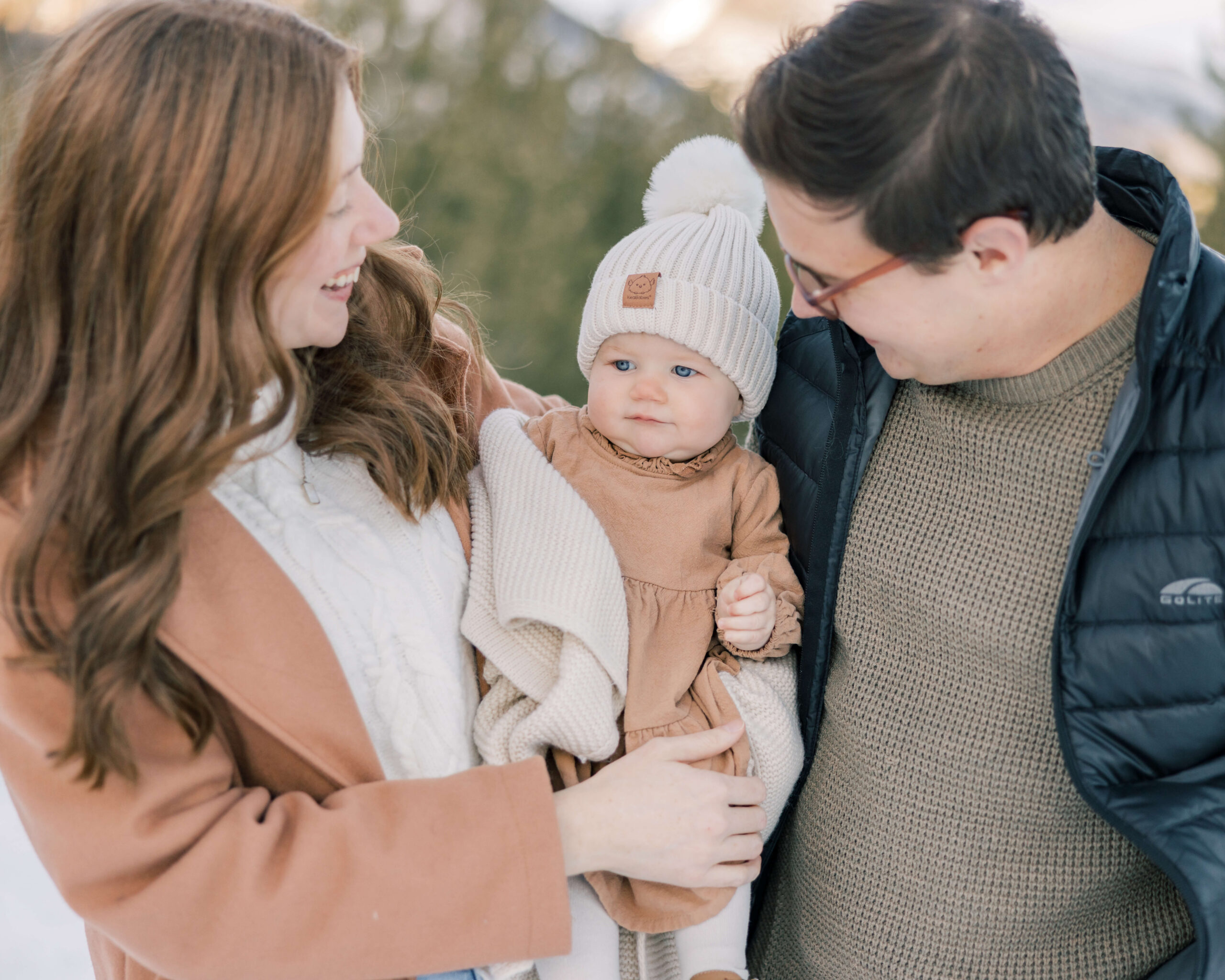 Mom and dad laugh with their toddler daughter between them while exploring the snow
