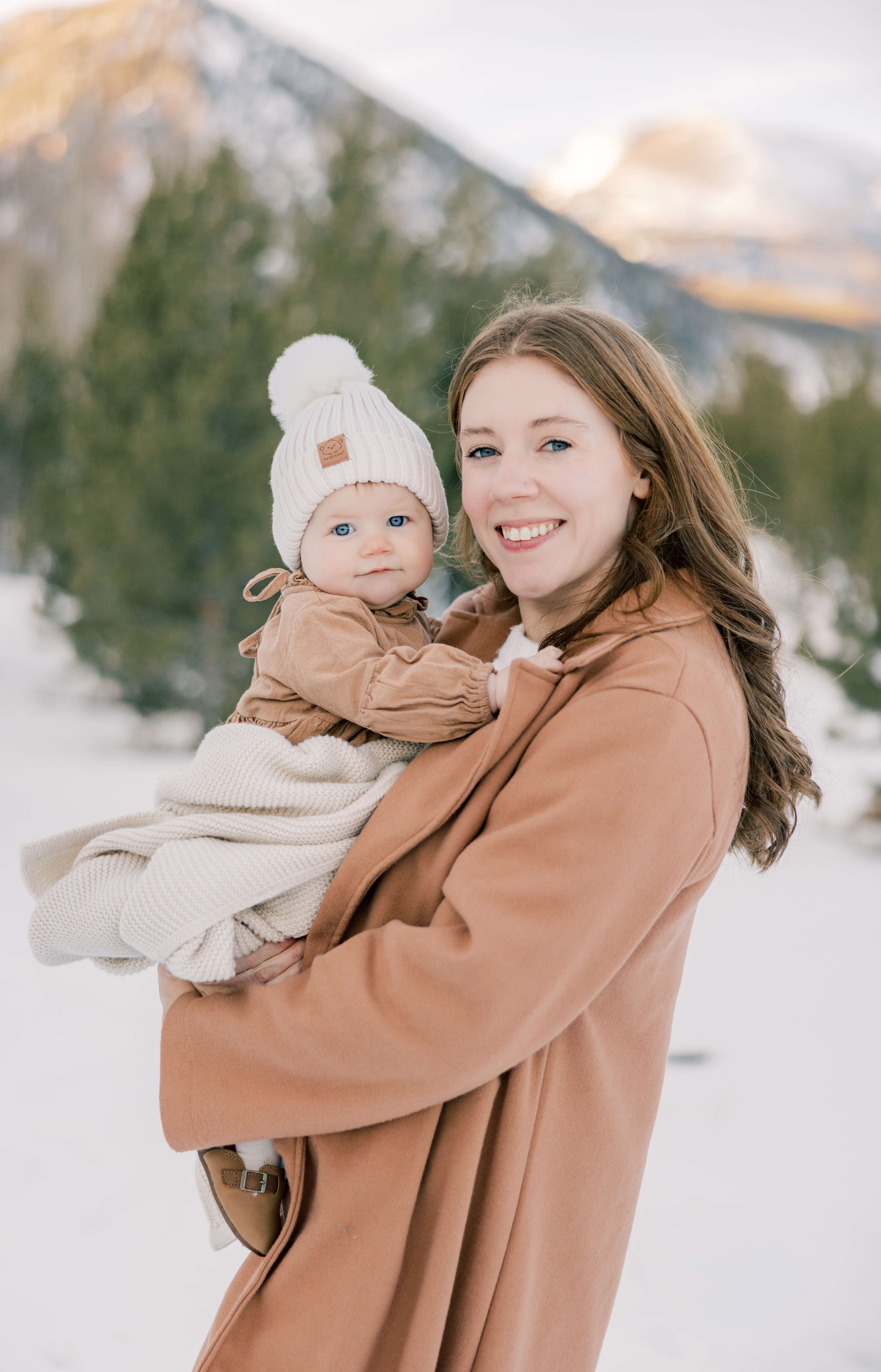 A happy mom in a brown coat holds her toddler daughter in a white beanie on a snow covered mountain at sunset after finding Breckenridge Childcare