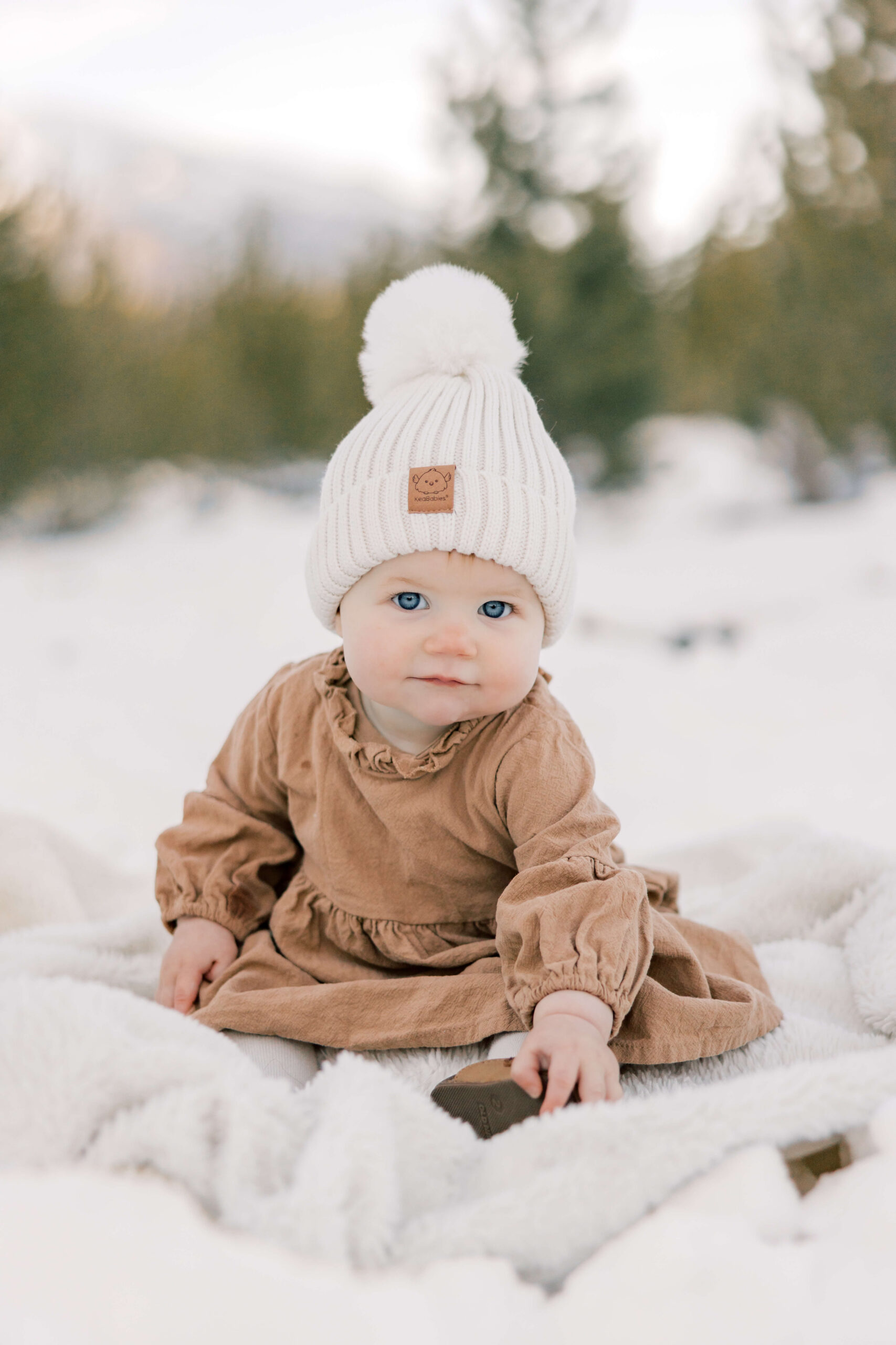 A baby girl in a brown dress and white beanie plays on a blanket in the snow at sunset after finding Breckenridge Childcare