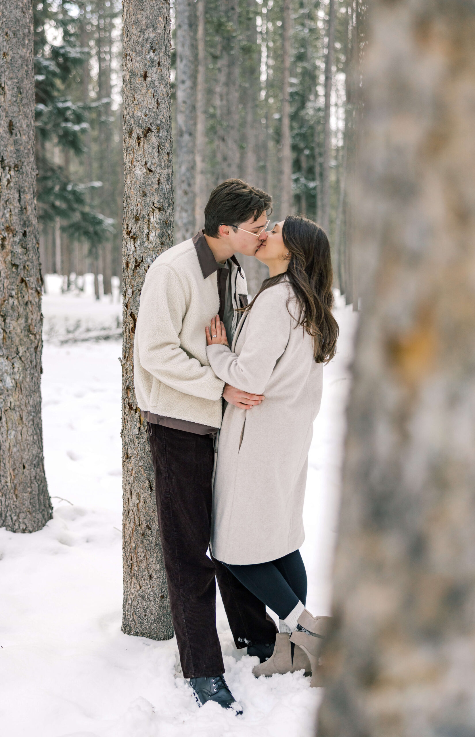 A happy couple in white sweaters enjoy a kiss against a tree in a snow covered forest