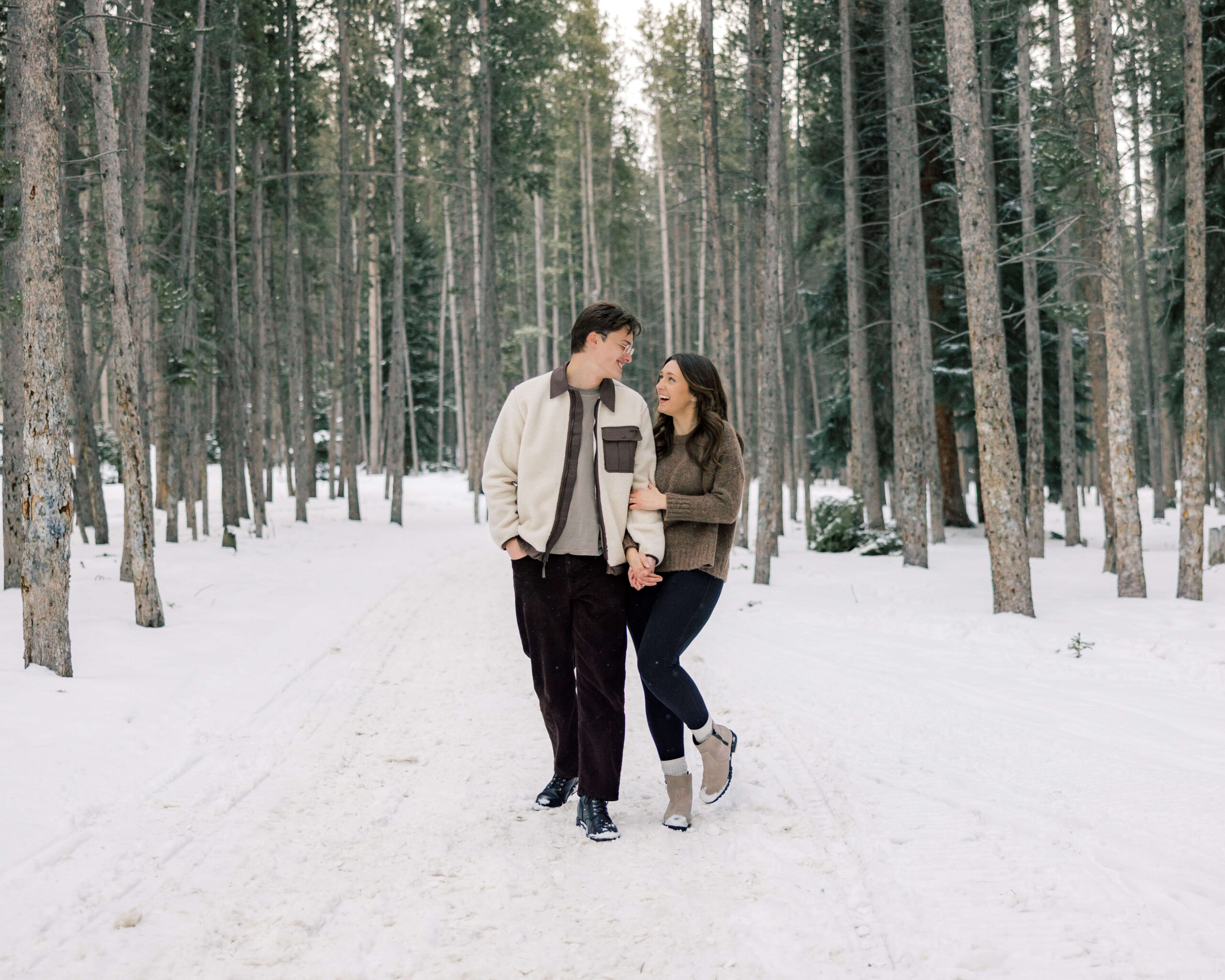 A laughing couple hike through a snow covered forest in sweaters and black pants during Romantic Things To Do In Breckenridge