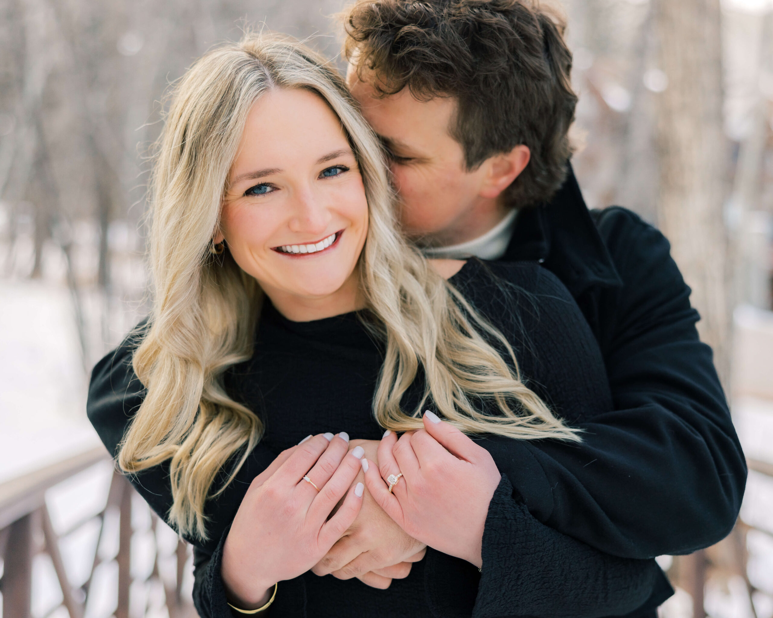 A smiling woman in a black sweater is hugged from behind by her fiancee on a pedestrian bridge in the snow after getting engaged