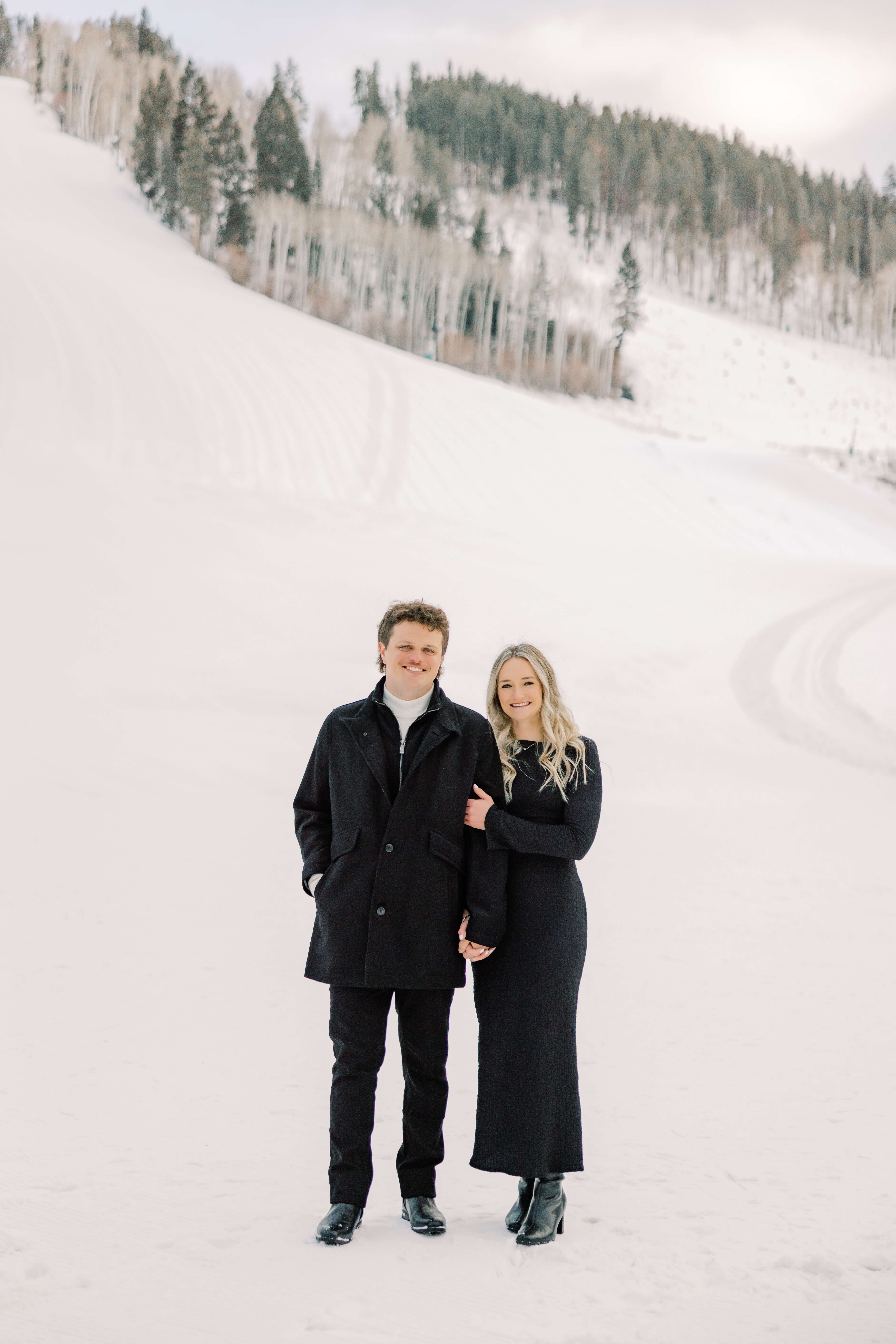 A smiling engaged couple stand holding hands in black outfits on a snow covered mountain after visiting luxury spas in Breckenridge
