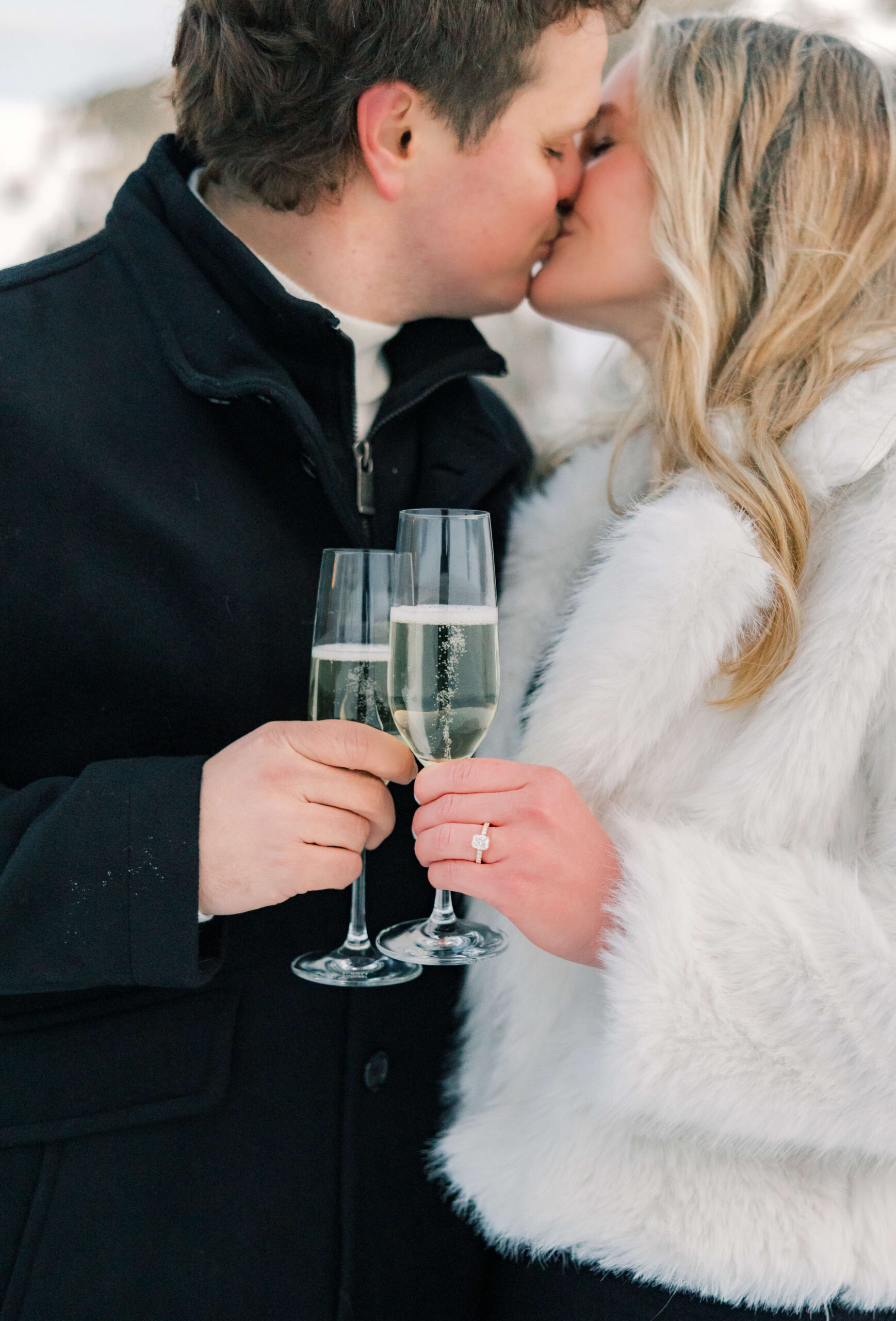 A happy newly engaged couple kisses while toasting champagne in black and white coats after visiting luxury spas in Breckenridge