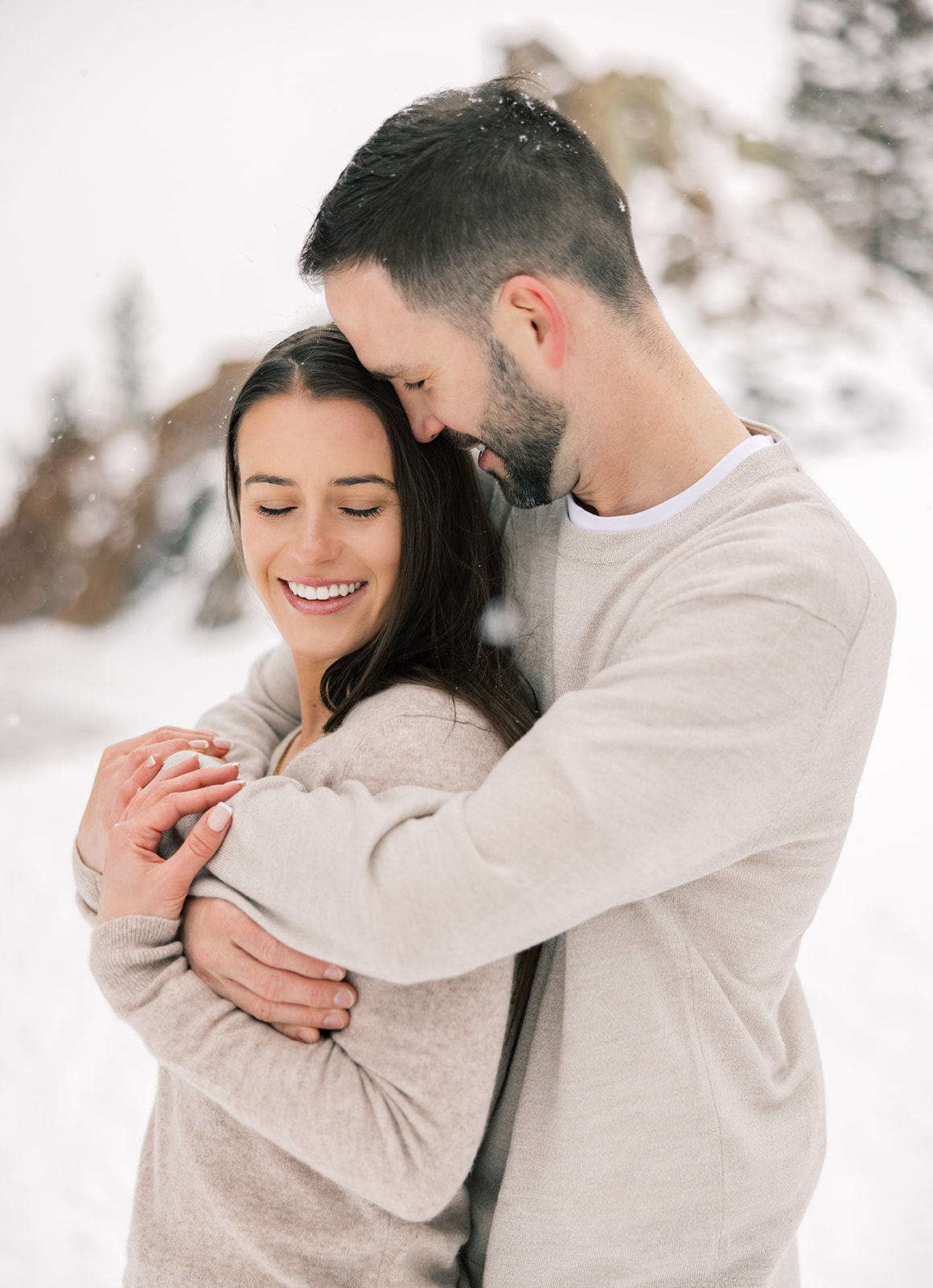 A woman snuggles into the chest of her fiancee both in tan sweaters as snow falls