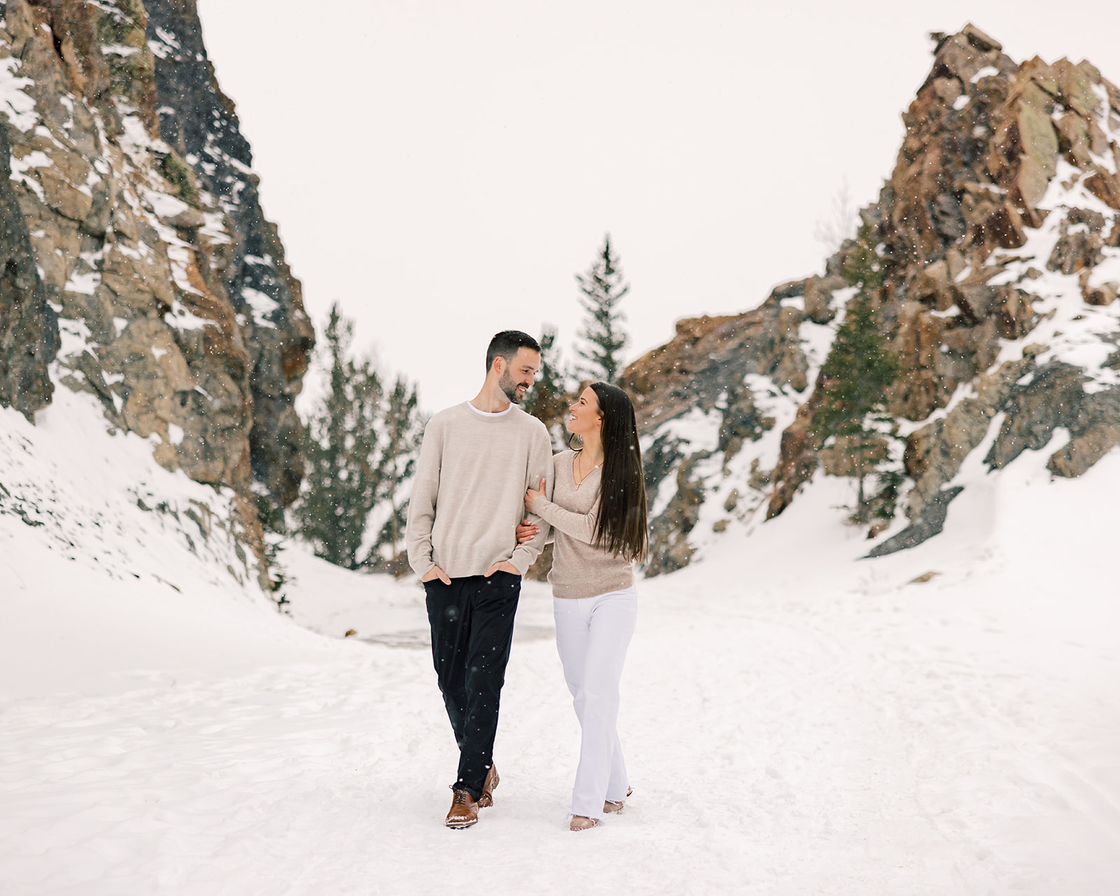 A happy engaged couple walk in a snowy mountain pass in tan sweaters after enjoying brunch in Breckenridge