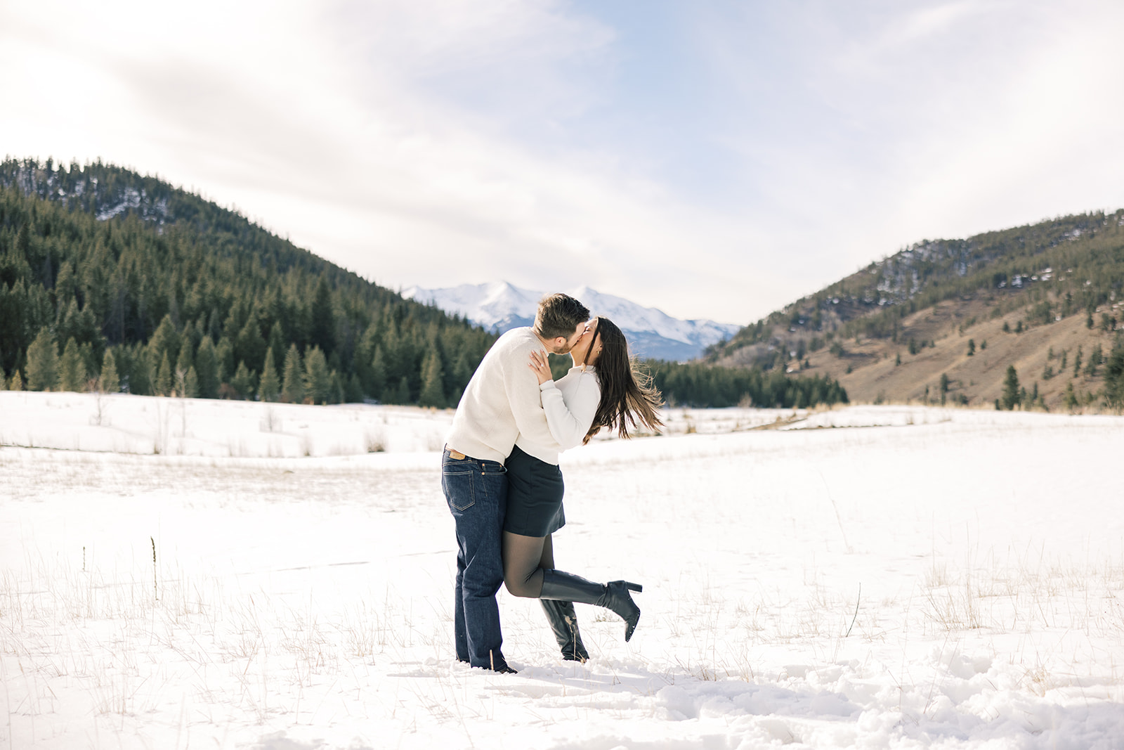 A newly engaged couple in white and denim kiss in a snow filled valley after one of the hot air balloon rides in Breckenridge, CO