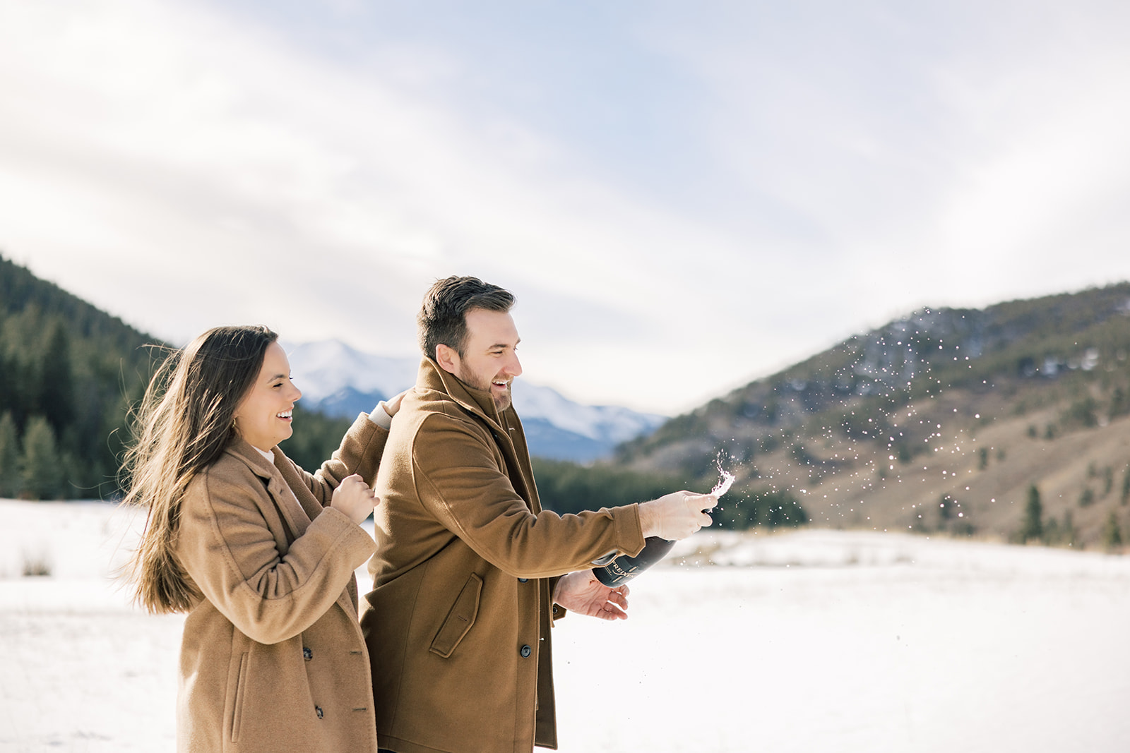 A happy couple in brown coats pop a bottle of champagne in a snow covered valley after one of the hot air balloon rides in Breckenridge, CO
