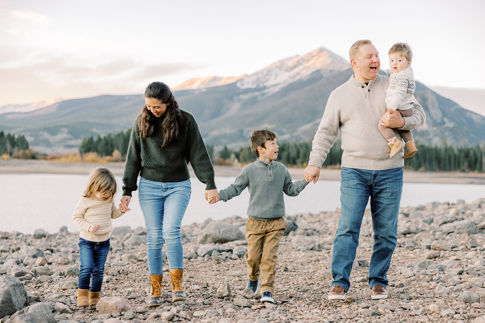 A mom and dad laugh with their three toddlers hiking by a lake in the mountains