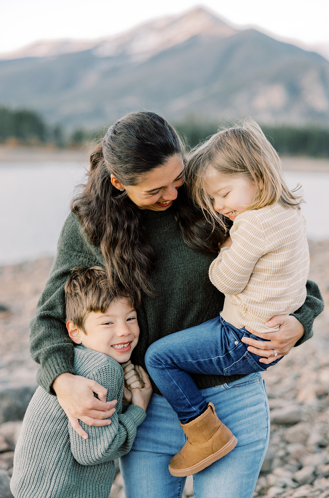 A mom giggles with her two toddlers as they snuggle by an alpine pond in sweaters and jeans before visiting peek-a-boo toys in Breckenridge