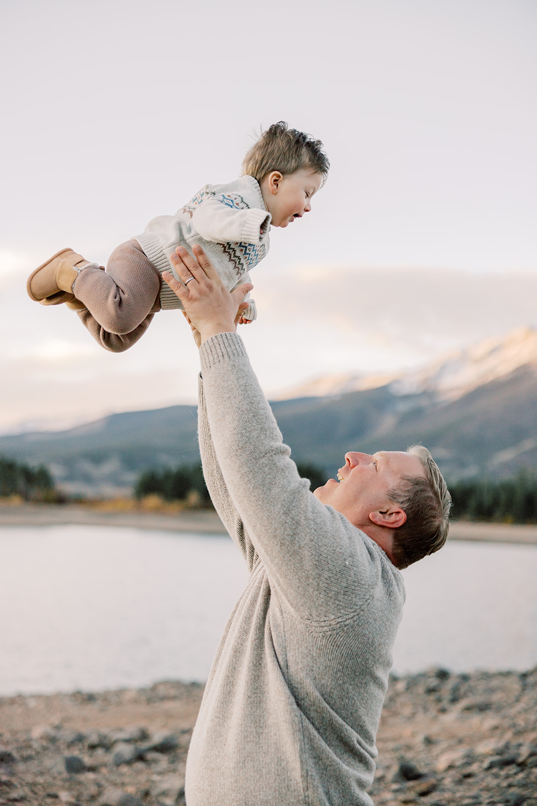 A happy dad tosses his toddler above his head by an alpine lake before visiting peek-a-boo toys in Breckenridge