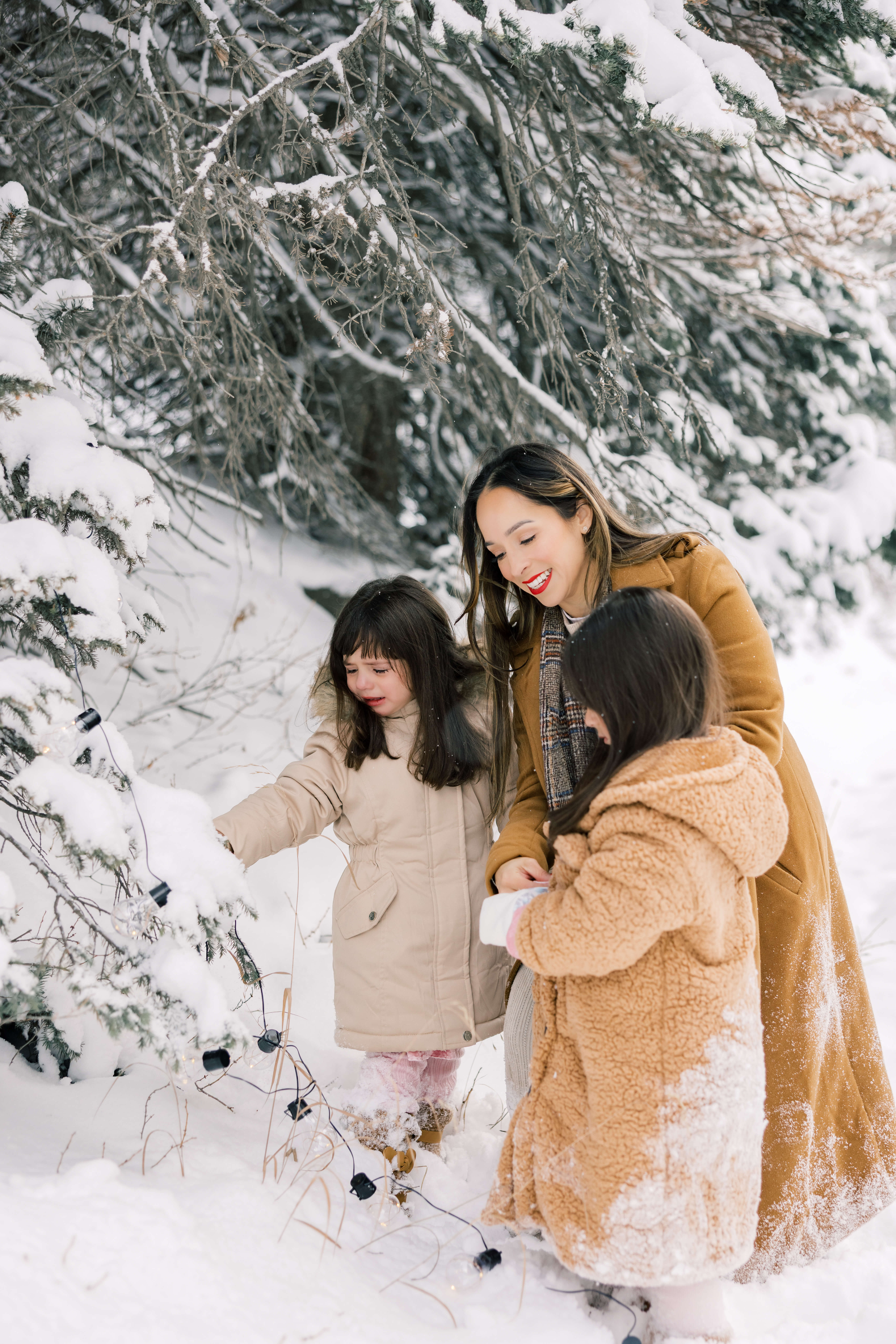 A mom helps her two toddler daughters hang lights on a tree in a snow covered forest after one of the Tours In Breckenridge