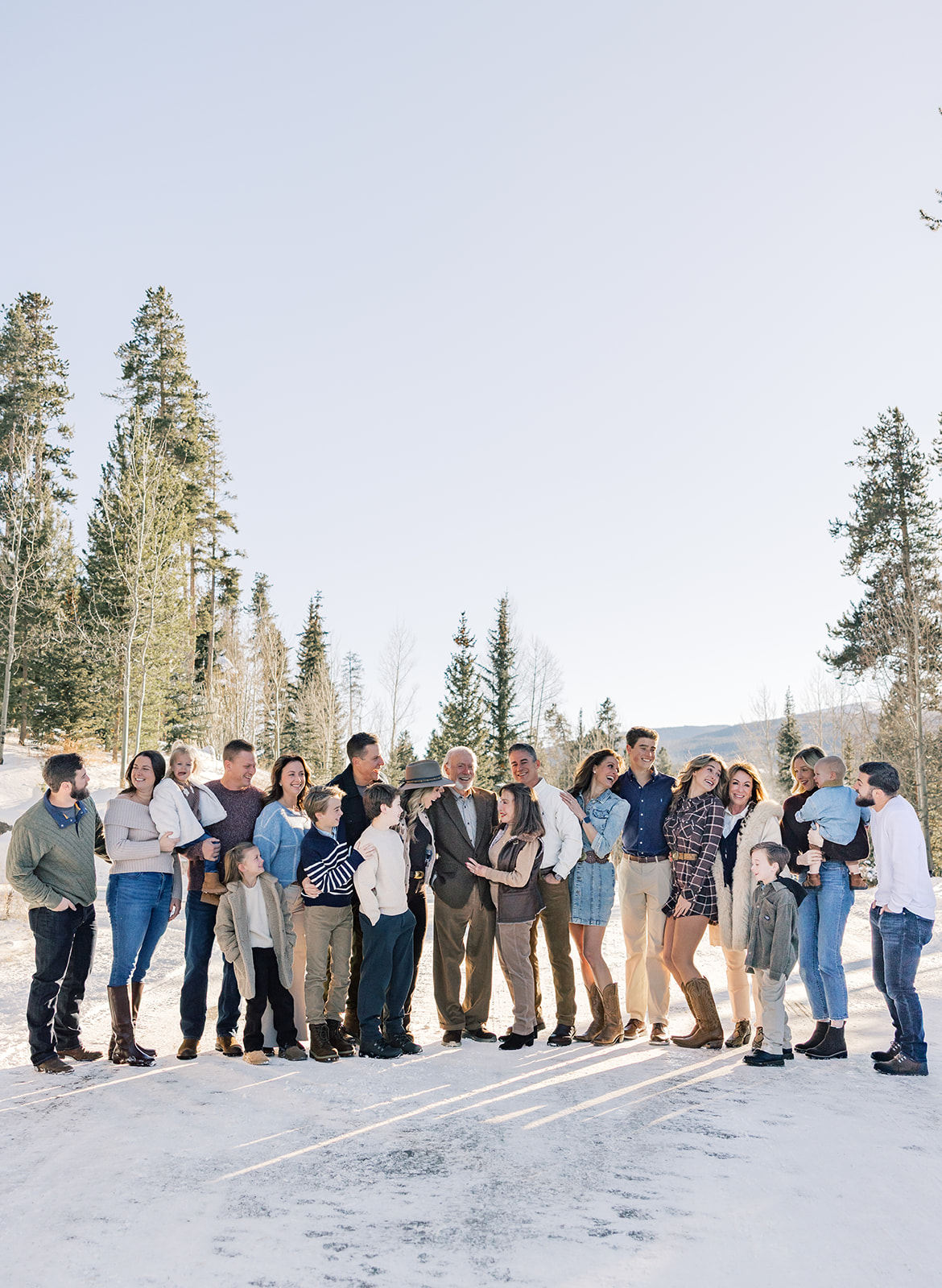 A large extended family smiles and laughs together in a snow covered meadow at sunset