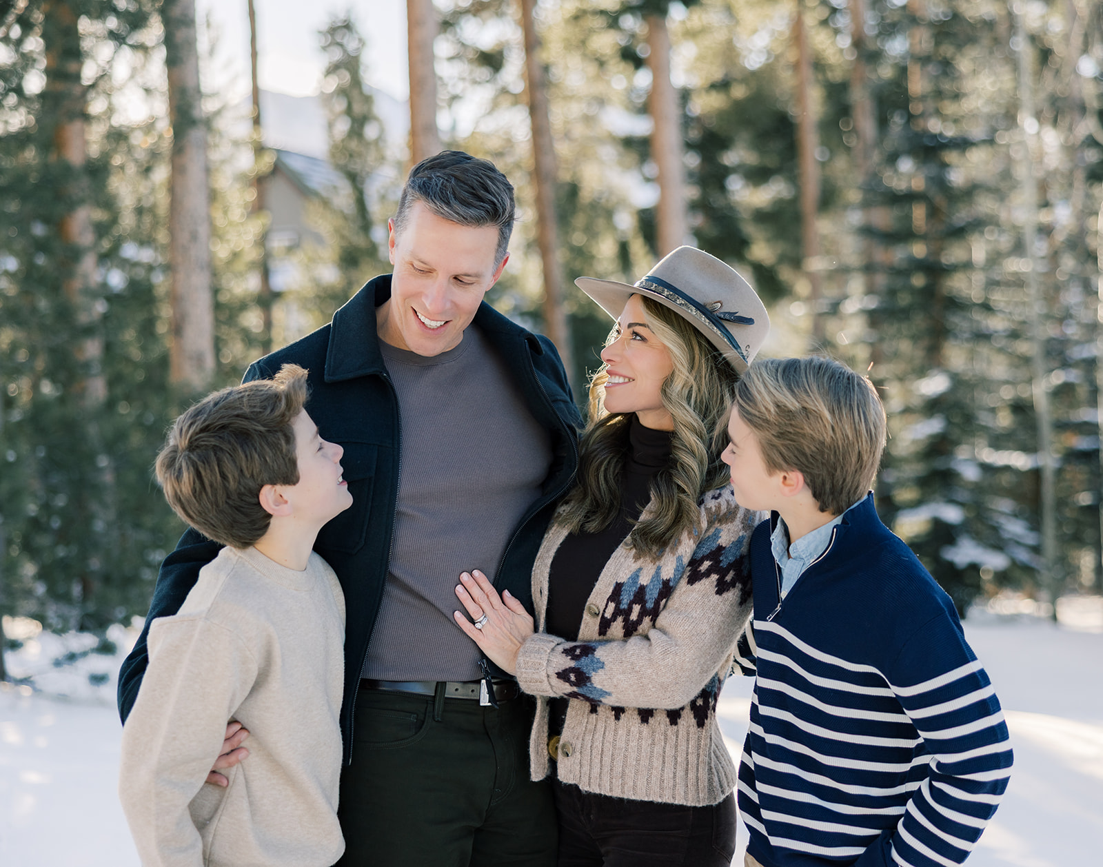 A happy family with two young boys laugh and stand in a snowy forest at sunset in sweaters after some outdoor ice skating in Breckenridge