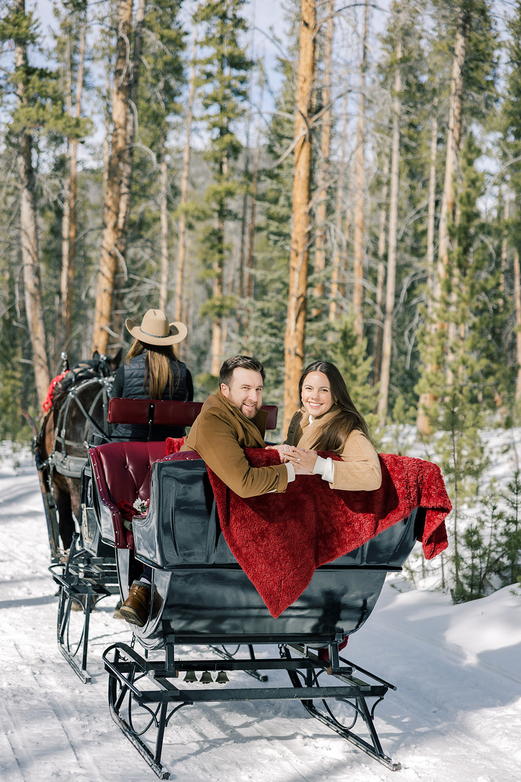 A happy couple sit in the back of a private sleigh ride in Breckenridge looking backwards with a red blanket holding hands and smiling