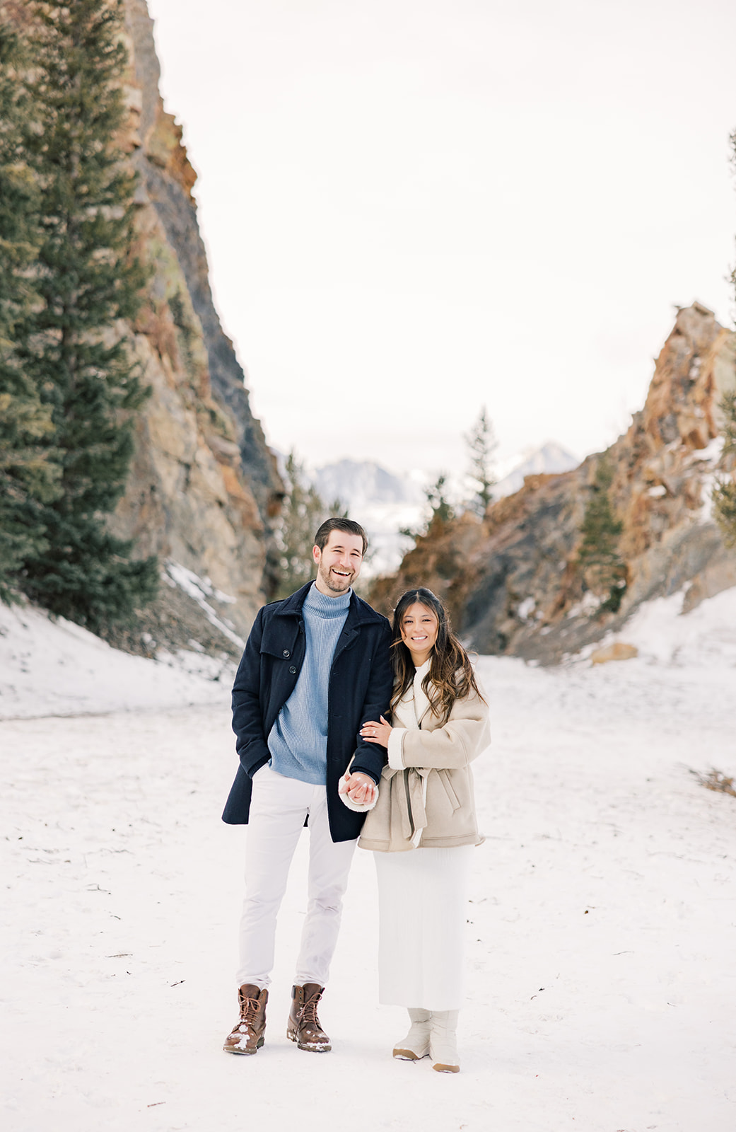 A happy couple walks holding hands while laughing in a snow covered mountain pass