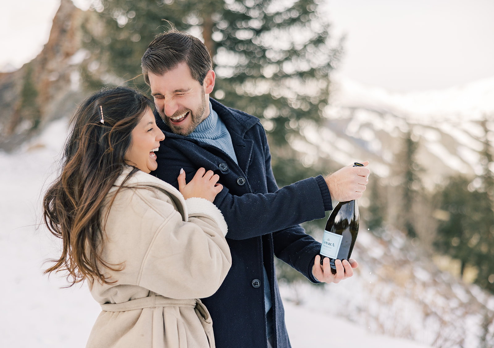 A happy couple in big jackets pop a bottle of champagne in a snow covered forest before enjoying things to do in Breckenridge at night