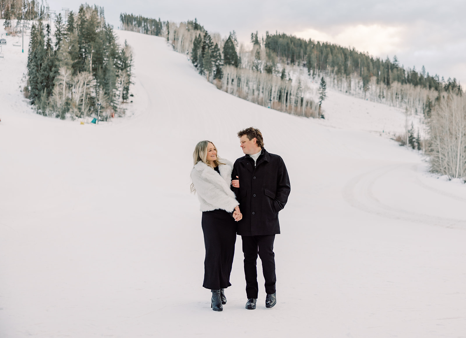 A happy engaged couple in black and white walk on a ski mountain holding hands and smiling at each other after finding great Breckenridge ski guides